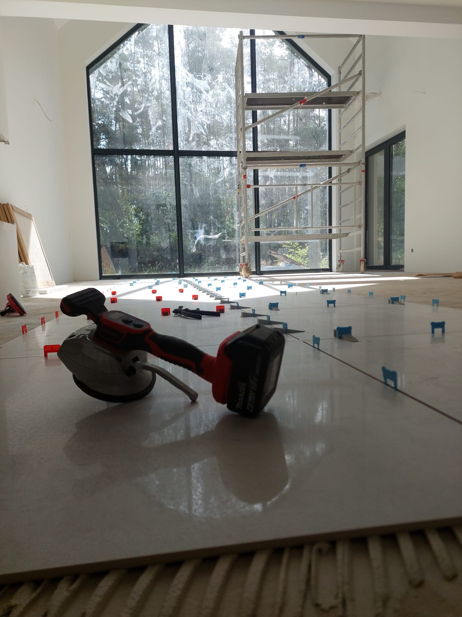 Układanie płytek podłogowych w jasnym wnętrzu z wysokim oknem. Widoczna poziomica wibracyjna Makita, krzyżyki dystansowe i rusztowanie. Praca w toku.