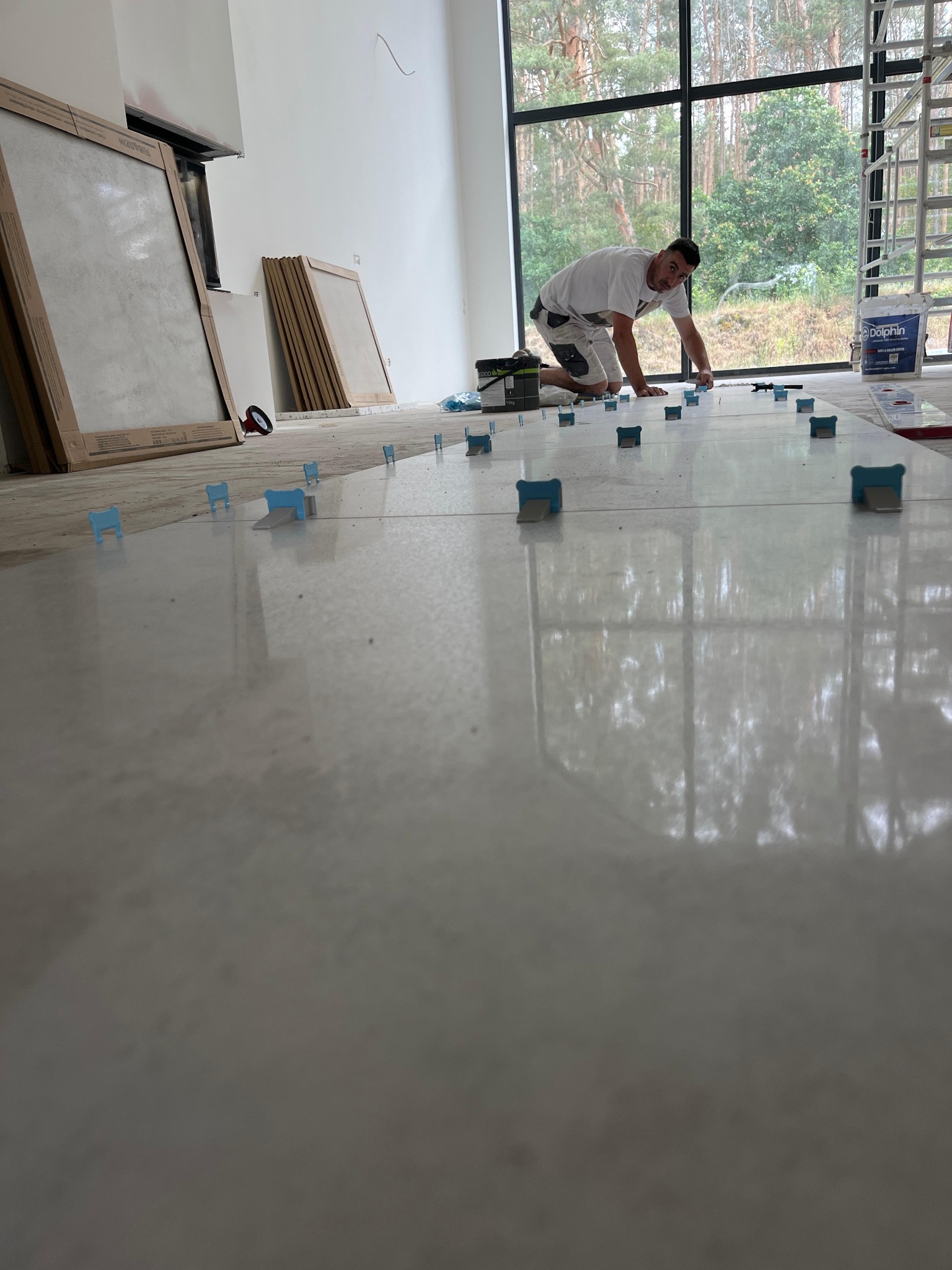 Glazurnik układa jasne płytki podłogowe, używając systemu poziomowania. Widoczne narzędzia i duże okno z widokiem na zieleń. Praca w toku, wnętrze w stanie surowym.