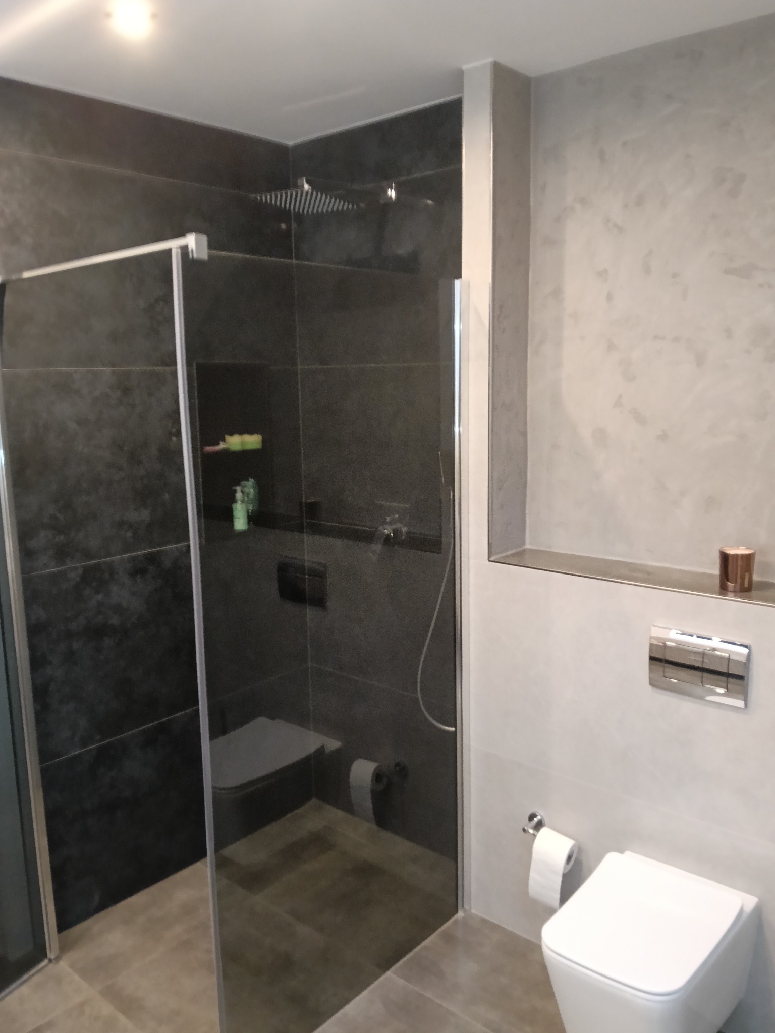 Nowoczesna łazienka z kabiną prysznicową typu walk-in, szare płytki i biała ceramika. Minimalistyczny design z wnęką i subtelnym oświetleniem.