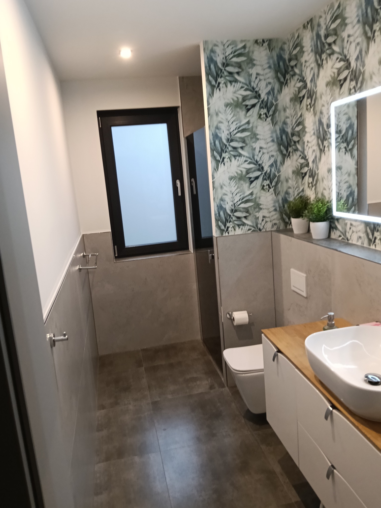 Nowoczesna łazienka z umywalką nablatową na drewnianym blacie, toaletą wiszącą i oknem. Ściana z wzorem liści, lustro z podświetleniem LED.