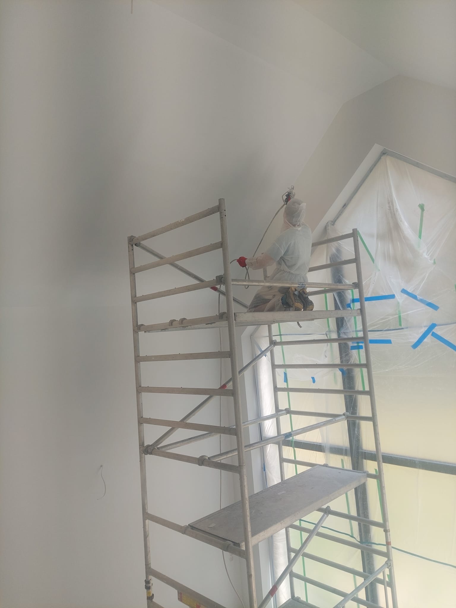 Pracownik w kombinezonie maluje ścianę natryskowo z rusztowania. Zabezpieczone okno folią malarską. Ściana w jasnym odcieniu szarości, widoczne niedoskonałości.