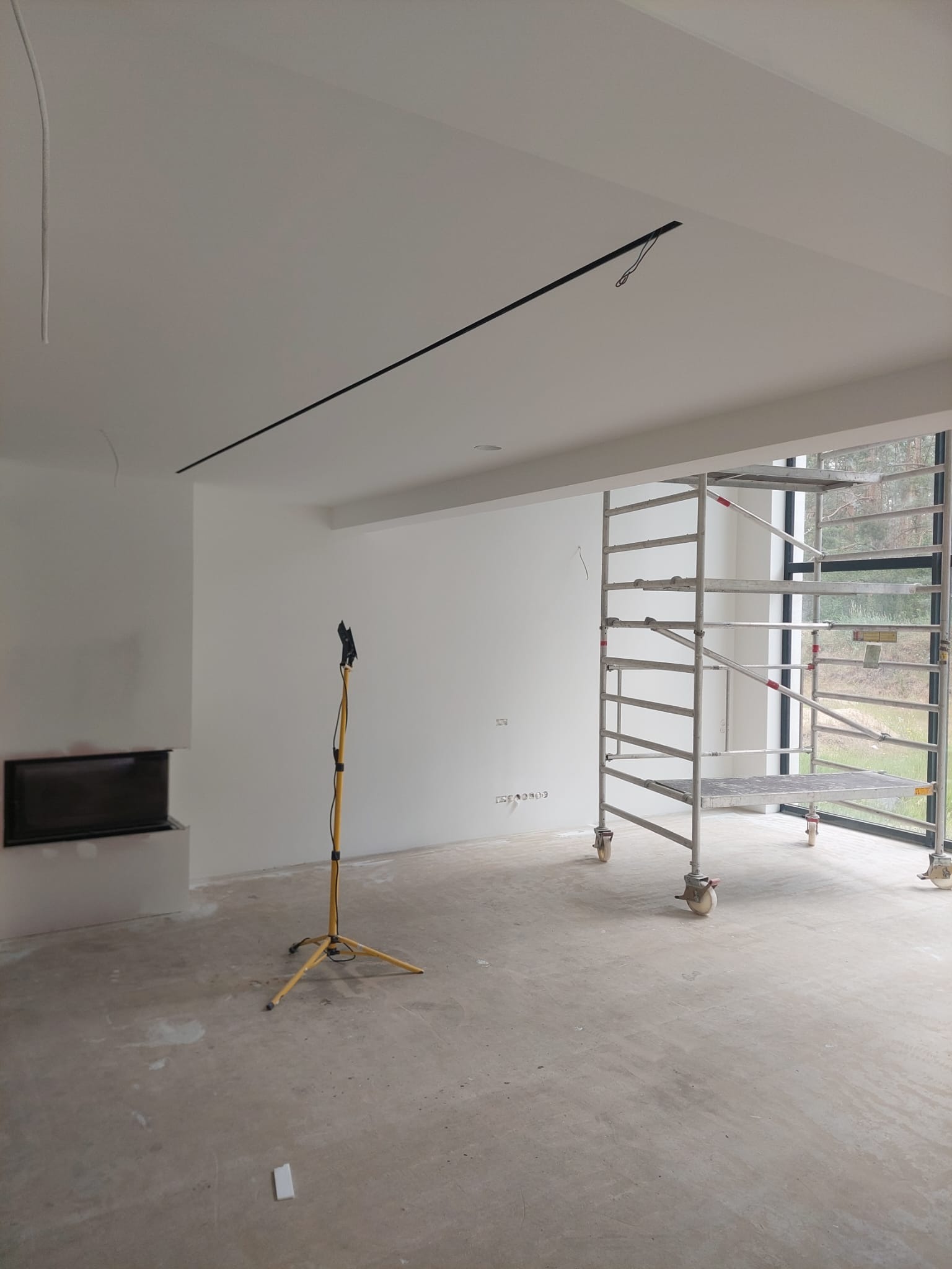 Wnętrze w trakcie prac wykończeniowych: białe ściany, surowa podłoga, rusztowanie przy oknie, lampa budowlana i czarny element dekoracyjny na suficie.