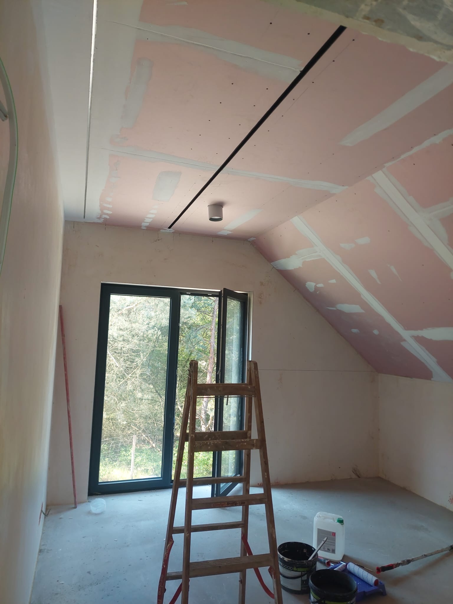 Poddasze w trakcie prac: płyty karton-gips na skosach, częściowo szpachlowane łączenia, drabina, okno z widokiem na zieleń, farby i wałki malarskie na podłodze.