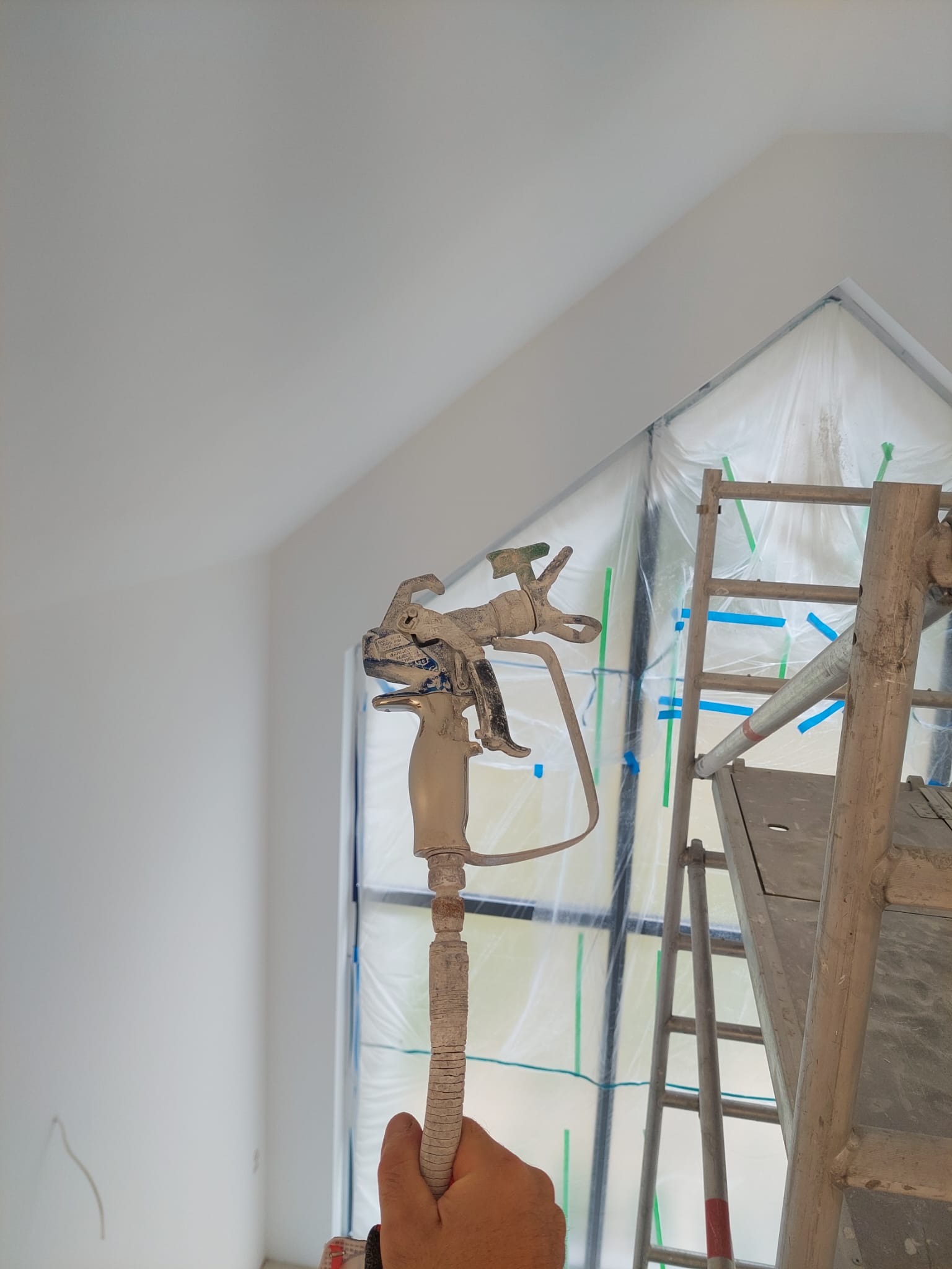 Malowanie natryskowe ścian w pokoju ze skosami. Widoczny pistolet natryskowy, drabina i okno zabezpieczone folią. Ściany świeżo pomalowane na biało.