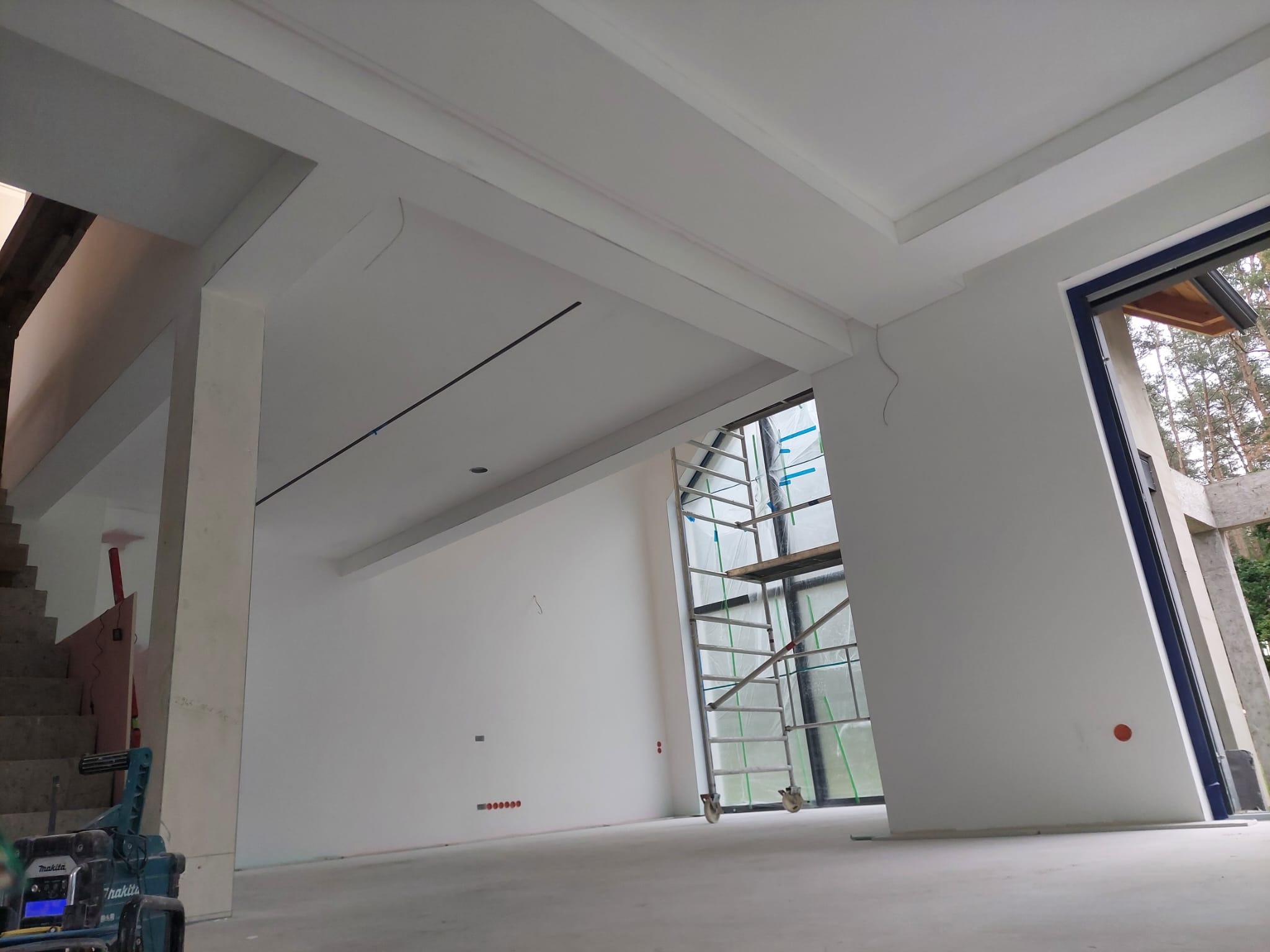 Wnętrze w trakcie wykończenia: białe ściany, sufit z dekoracyjnymi listwami, rusztowanie przy oknie, widoczne instalacje elektryczne. Surowy stan, przygotowanie do malowania.