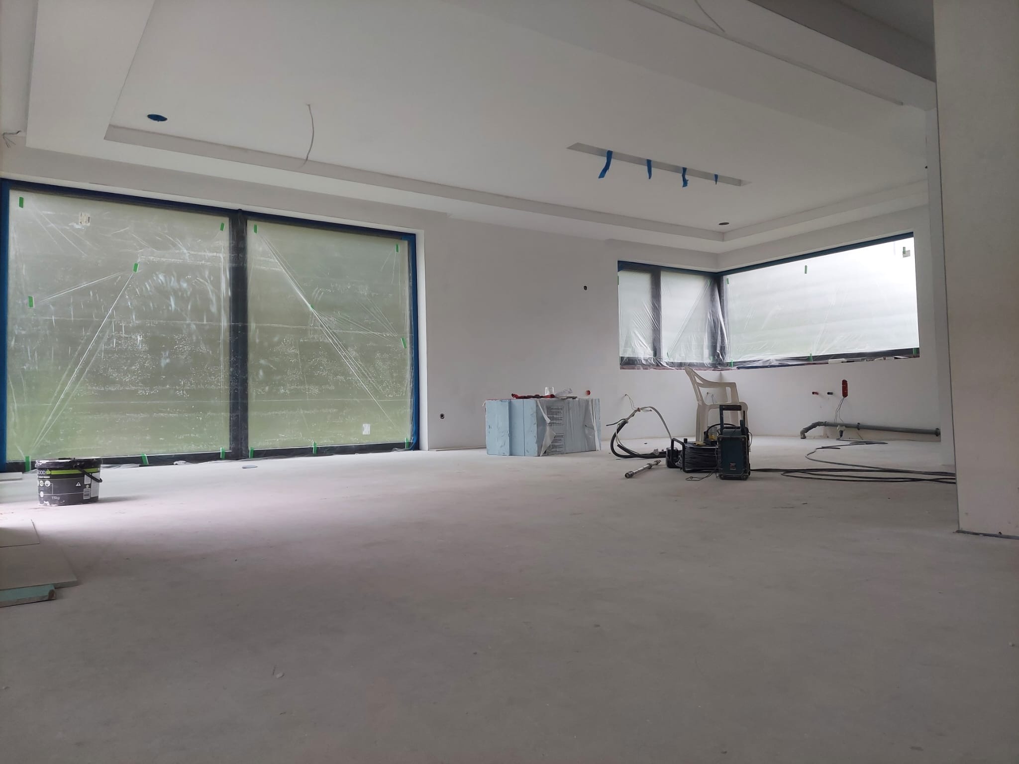 Wnętrze w trakcie prac wykończeniowych: zabezpieczone okna folią, świeżo wylana posadzka, widoczne narzędzia i materiały budowlane. Przygotowanie do dalszych prac.