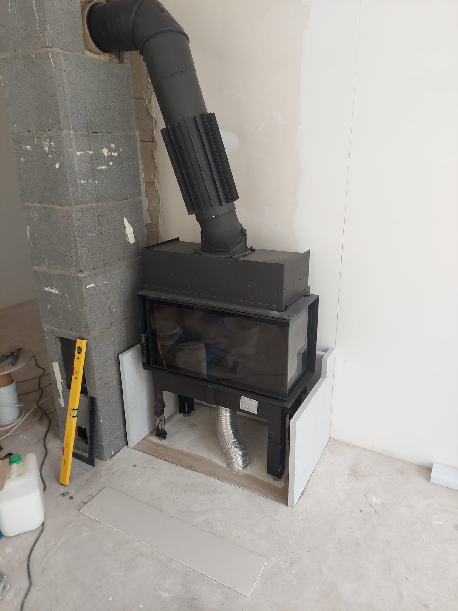 Montaż wkładu kominkowego z czarnym przewodem dymowym w surowym wnętrzu. Widoczny bloczek betonowy, poziomica i płyta gipsowa. Etap prac wykończeniowych.