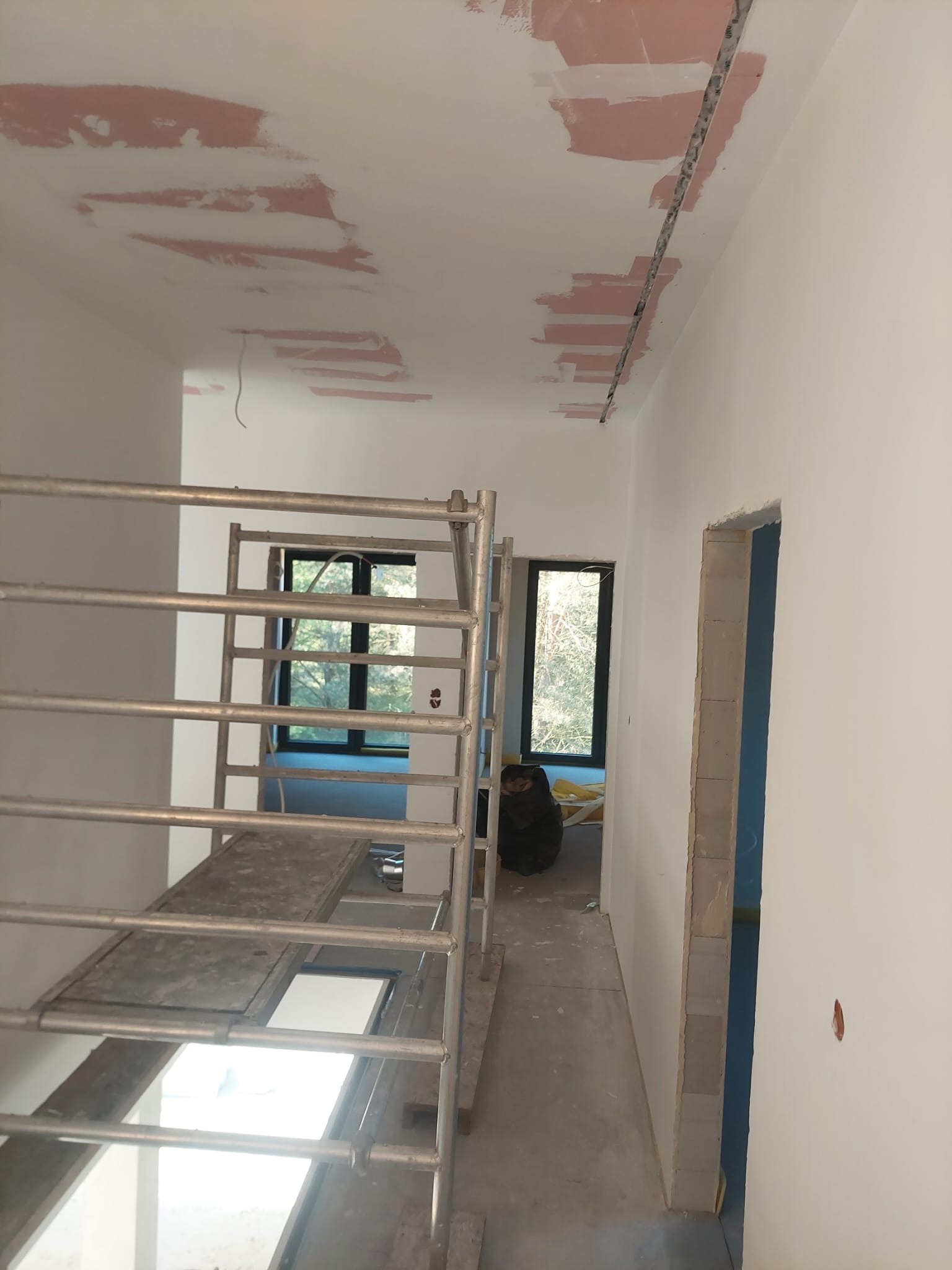 Wnętrze w trakcie prac wykończeniowych: szpachlowana sufit z widocznymi łatami, metalowe rusztowanie, przygotowane do malowania ściany i futryny drzwiowe.