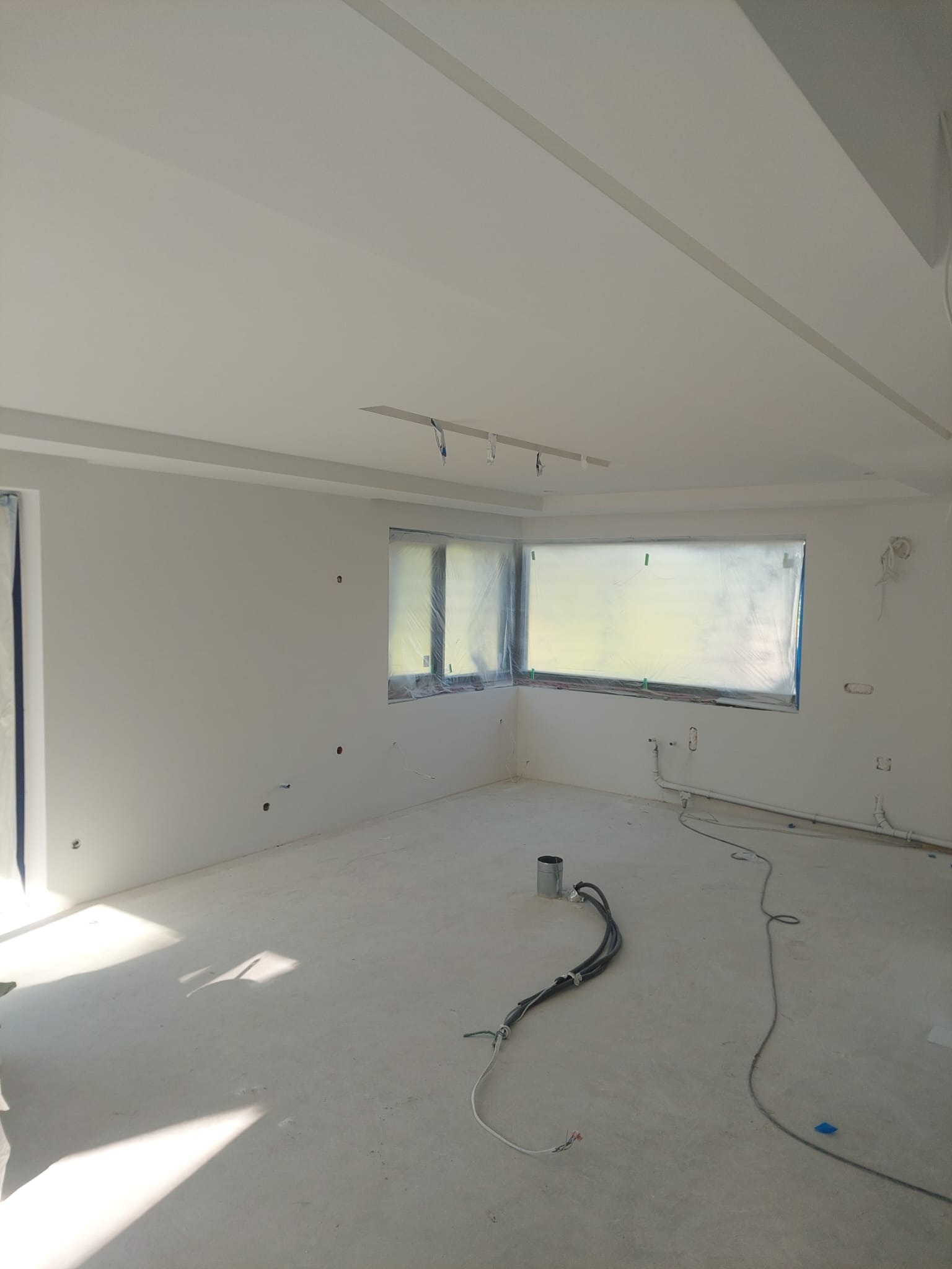 Jasne wnętrze w trakcie prac wykończeniowych: świeżo pomalowane ściany, zabezpieczone okna folią, widoczne przewody elektryczne i rury na podłodze. Minimalistyczny sufit.