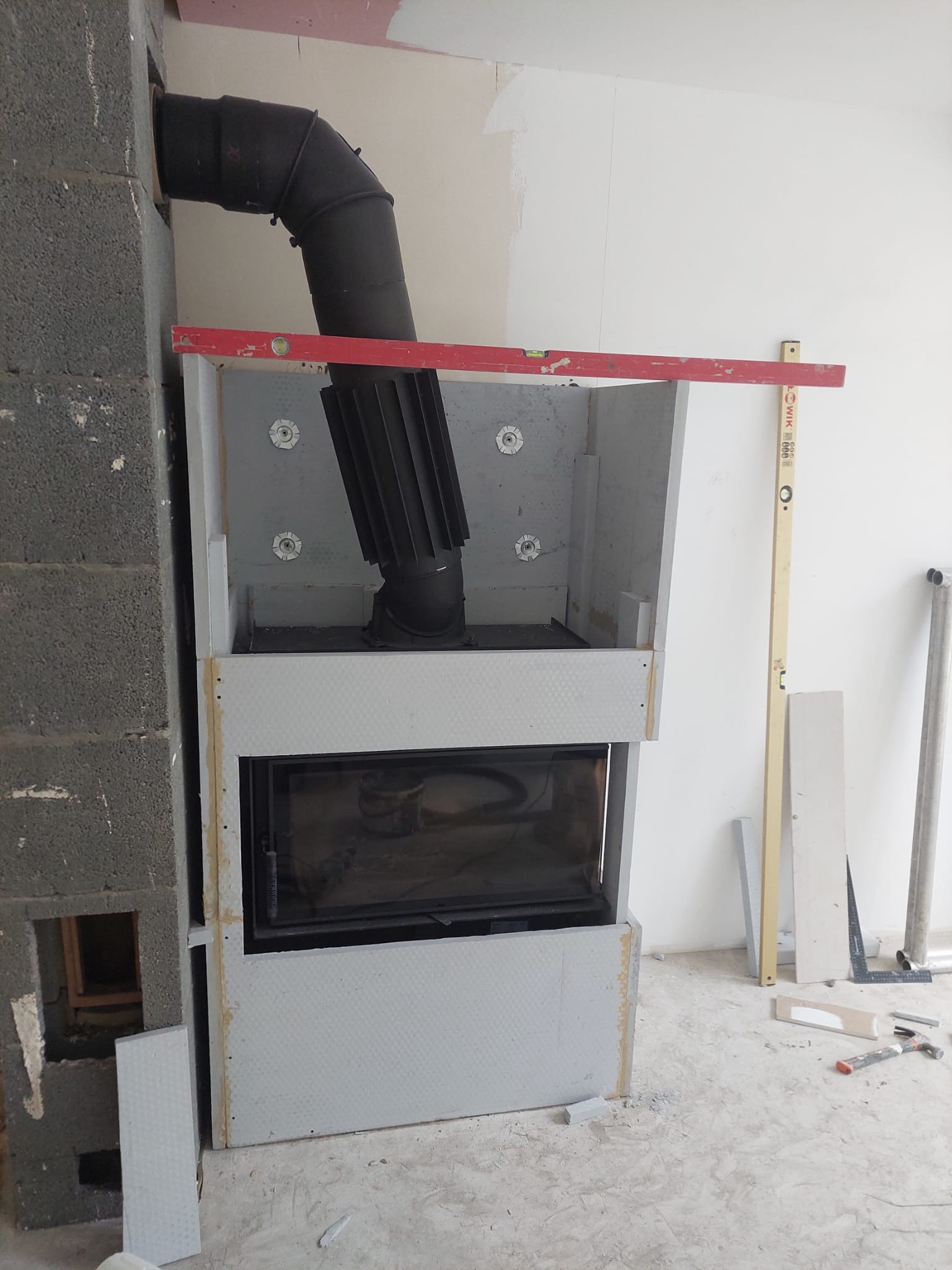 Montaż wkładu kominkowego z obudową w surowym stanie. Widoczne narzędzia i poziomica. Fragment ściany z pustaków. Prace wykończeniowe w trakcie.