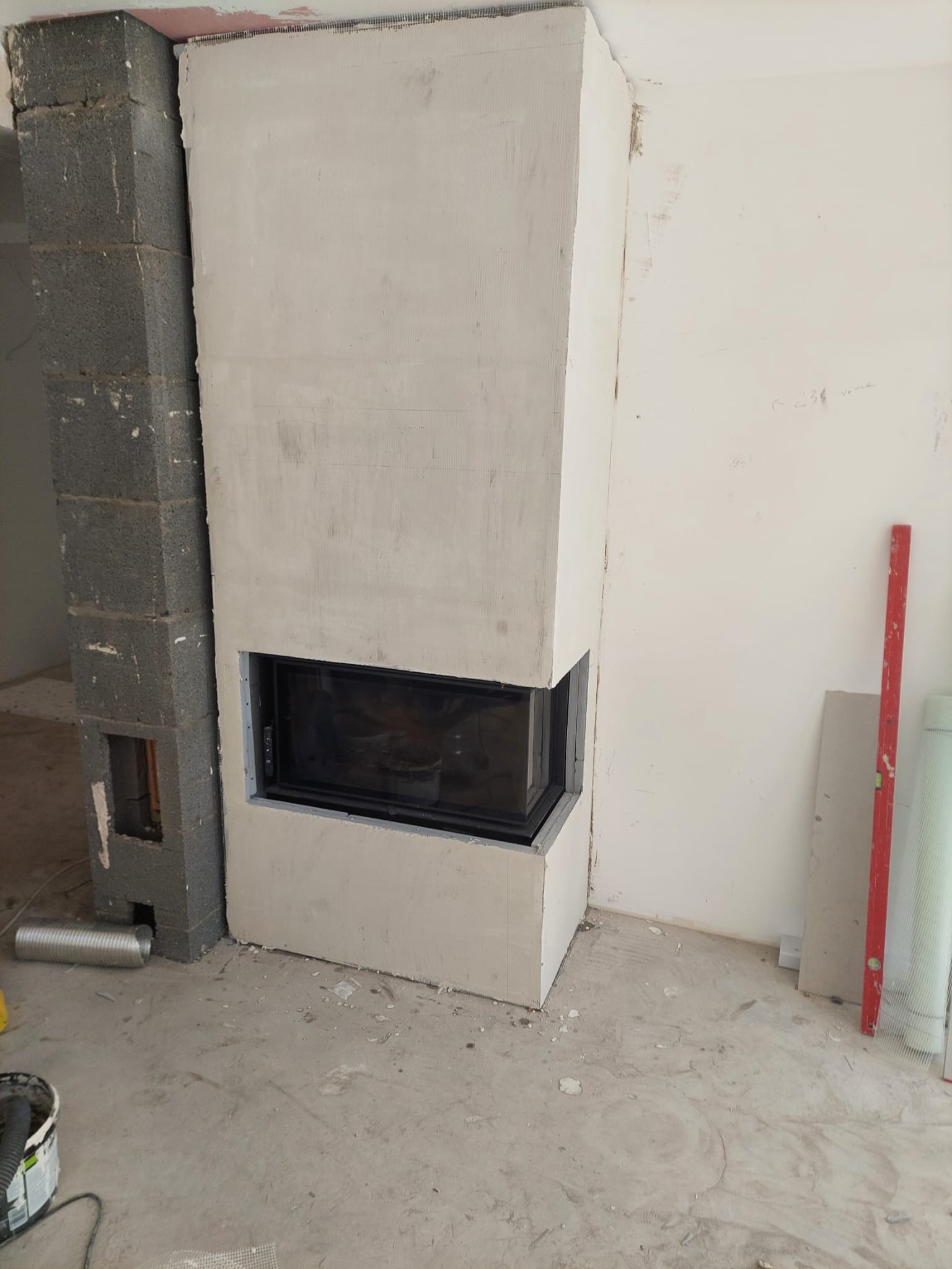 Wnętrze w trakcie wykańczania z obudowanym kominkiem z czarnym wkładem, widoczny fragment ściany z bloczków i narzędzia budowlane na podłodze.