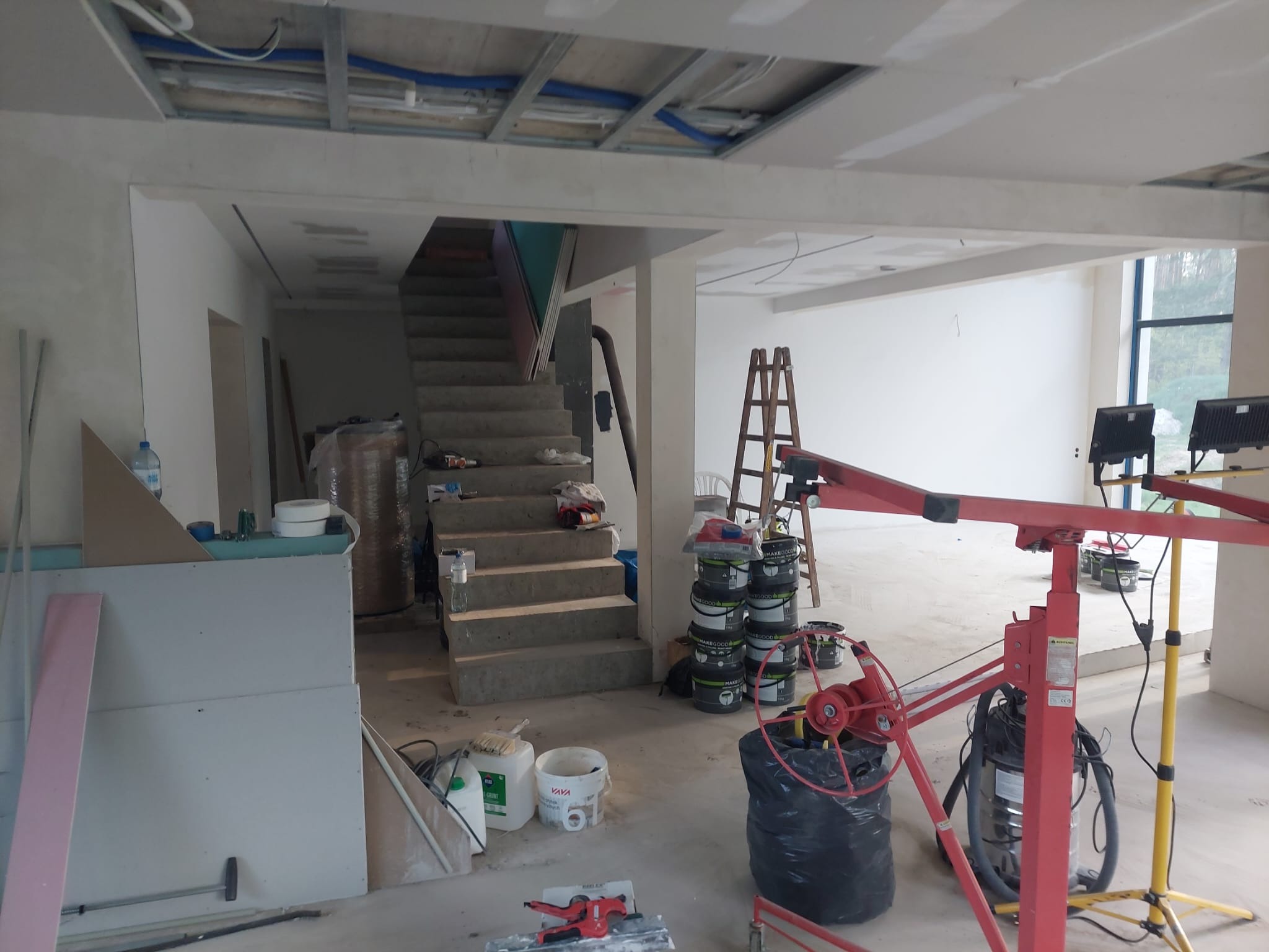 Wnętrze w trakcie prac wykończeniowych: surowy strop z instalacjami, ściany przygotowane do malowania, narzędzia i materiały budowlane. Widoczne schody i podnośnik do płyt.