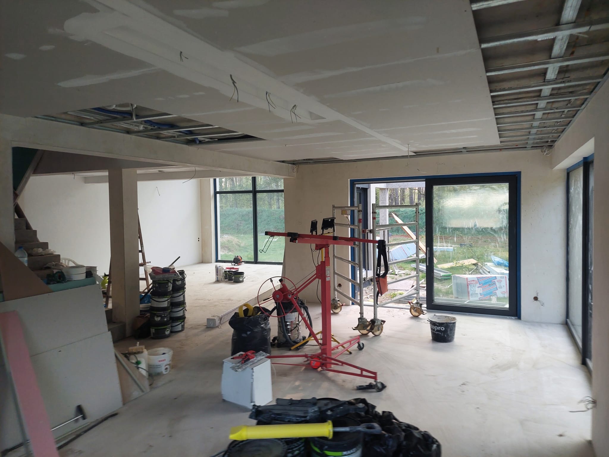 Montaż płyt kartonowo-gipsowych na suficie w przestronnym wnętrzu. Widoczny podnośnik do płyt, metalowa konstrukcja sufitu i częściowo wykończone ściany.