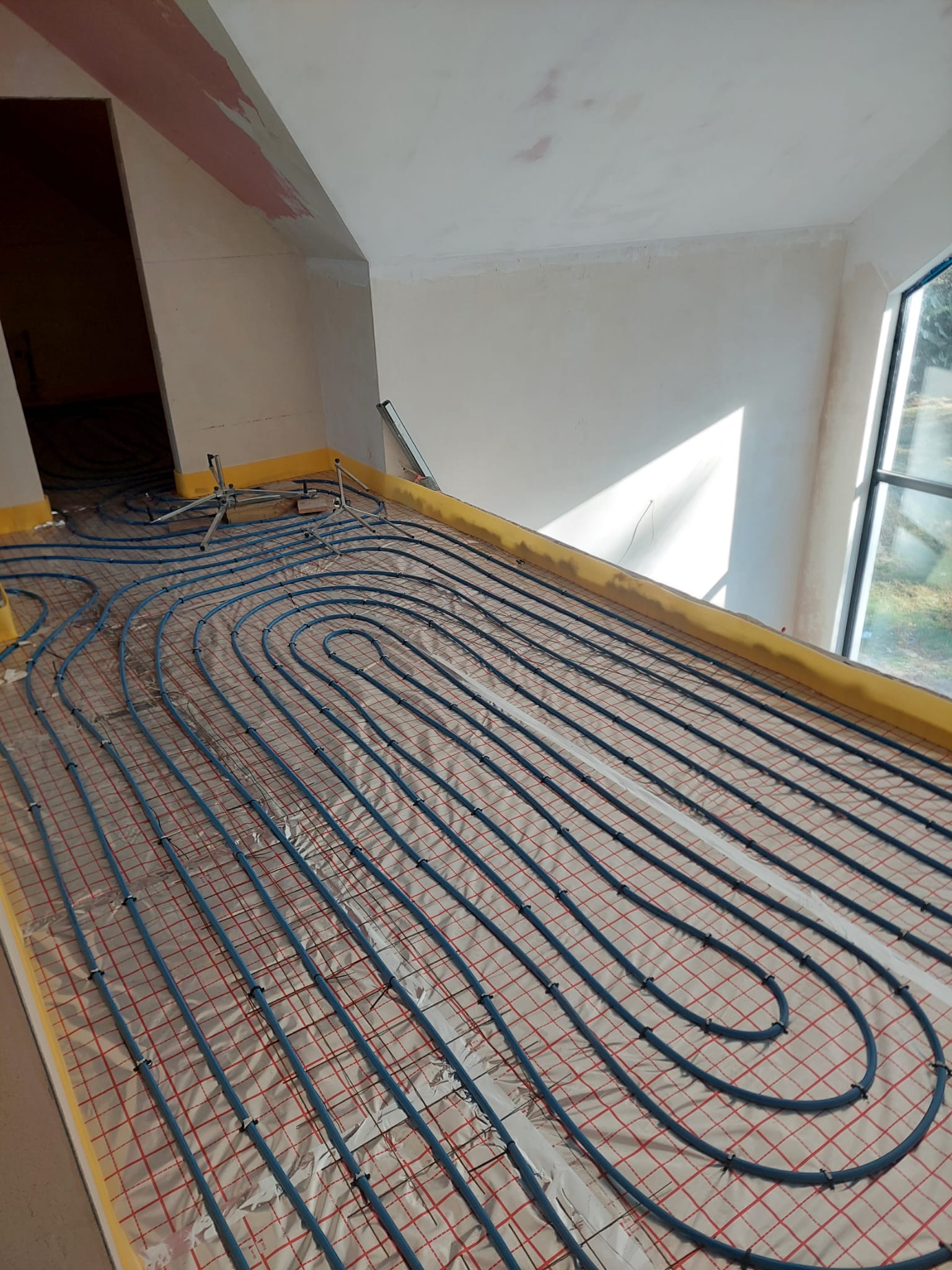 Niebieskie rury ogrzewania podłogowego ułożone na folii z czerwoną kratką w jasnym pomieszczeniu z oknem. Widoczne fragmenty ścian i sufitu w trakcie prac wykończeniowych.