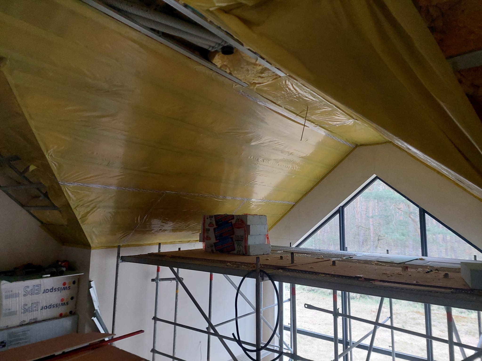 Ocieplenie poddasza folią i wełną mineralną, widok z rusztowania. Okno trójkątne w szczycie dachu. Częściowo widoczne materiały izolacyjne i narzędzia.