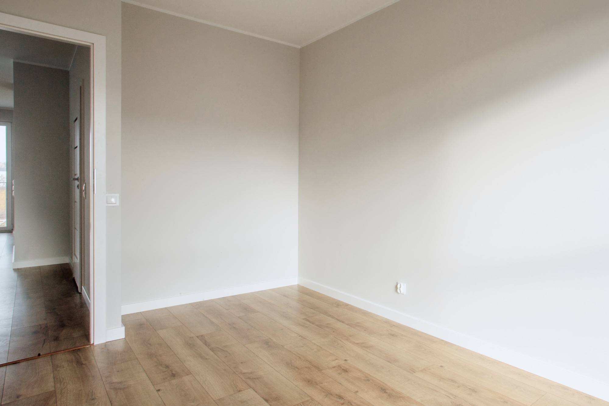Jasne wnętrze po remoncie: świeżo pomalowane ściany w odcieniach szarości, panele podłogowe imitujące drewno, białe listwy przypodłogowe i widok na kolejne pomieszczenie przez otwarte drzwi.
