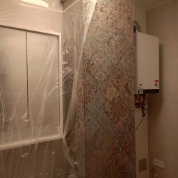 LUK-BUD Usługi Ogólnobudowlane Łukasz Barański - Wykończona ściana w łazience z dekoracyjnymi płytkami, zabezpieczona folią wanna i piec gazowy. Widoczne gniazdka elektryczne i wentylacja.