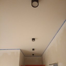 LUK-BUD Usługi Ogólnobudowlane Łukasz Barański - Przygotowane do malowania wnętrze z taśmą malarską na styku ścian i sufitu, widoczne czarne, okrągłe lampy sufitowe w rzędzie. Ściany w trakcie prac.