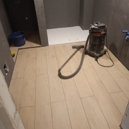 LUK-BUD Usługi Ogólnobudowlane Łukasz Barański - Jasne panele podłogowe w łazience z szarą ścianą i brodzikiem. Odkurzacz przemysłowy stoi na świeżo ułożonej podłodze. Widoczne niedokończone wykończenia.
