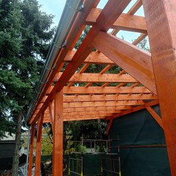 LUK-BUD Usługi Ogólnobudowlane Łukasz Barański - Drewniana konstrukcja z orynnowaniem, widok z dołu. Świeżo zaimpregnowane drewno w ciepłym odcieniu brązu. W tle drzewa i rusztowanie.