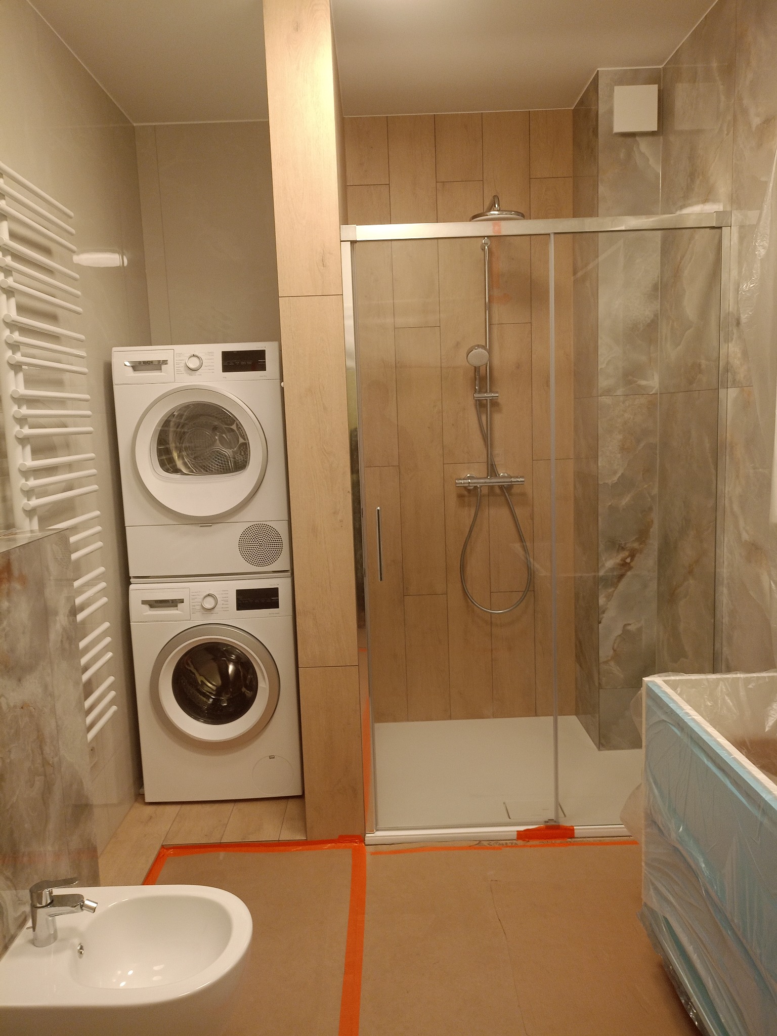 Wykończona łazienka z kabiną prysznicową, pralką i suszarką w kolumnie. Beżowe płytki imitujące drewno i marmur. Bidet na pierwszym planie.