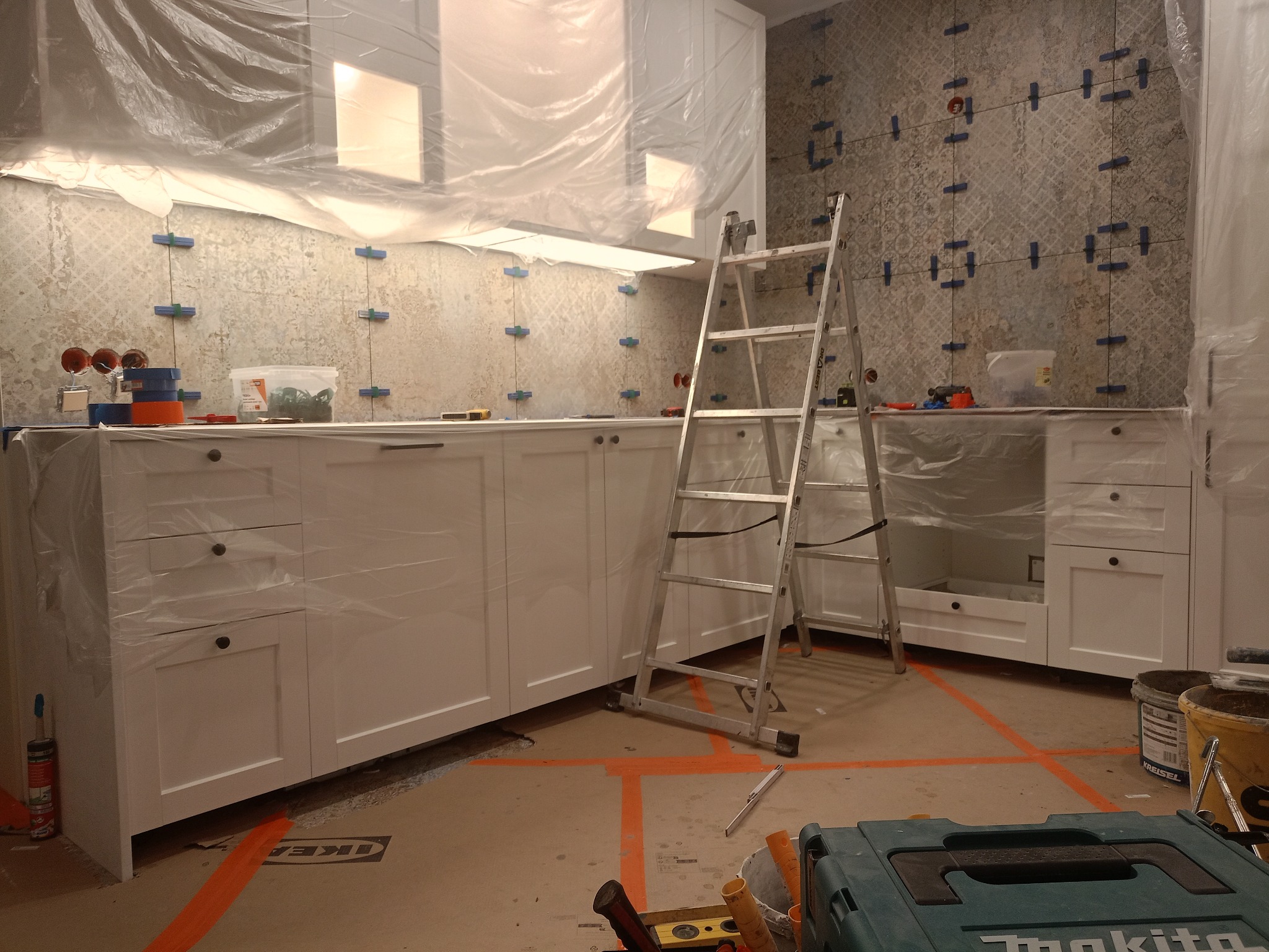 Układanie płytek w kuchni w Mechlinie. Widoczne białe szafki, drabina i narzędzia. Ściana w trakcie prac, z niebieskimi krzyżykami dystansowymi. Folia ochronna na meblach.