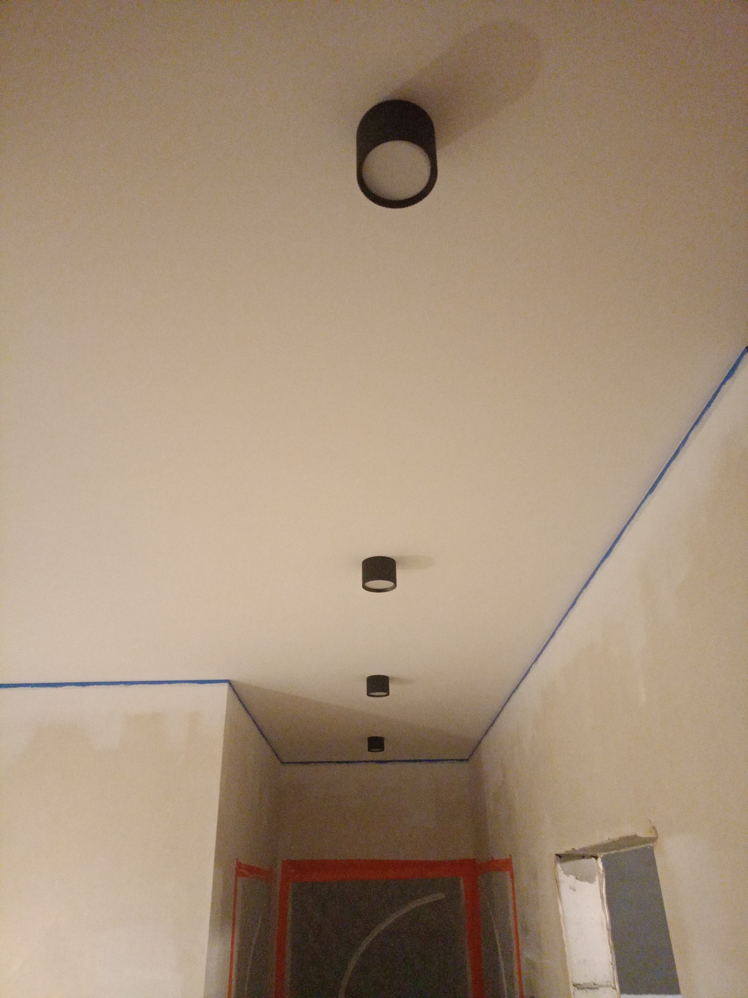Przygotowane do malowania wnętrze z taśmą malarską na styku ścian i sufitu, widoczne czarne, okrągłe lampy sufitowe w rzędzie. Ściany w trakcie prac.