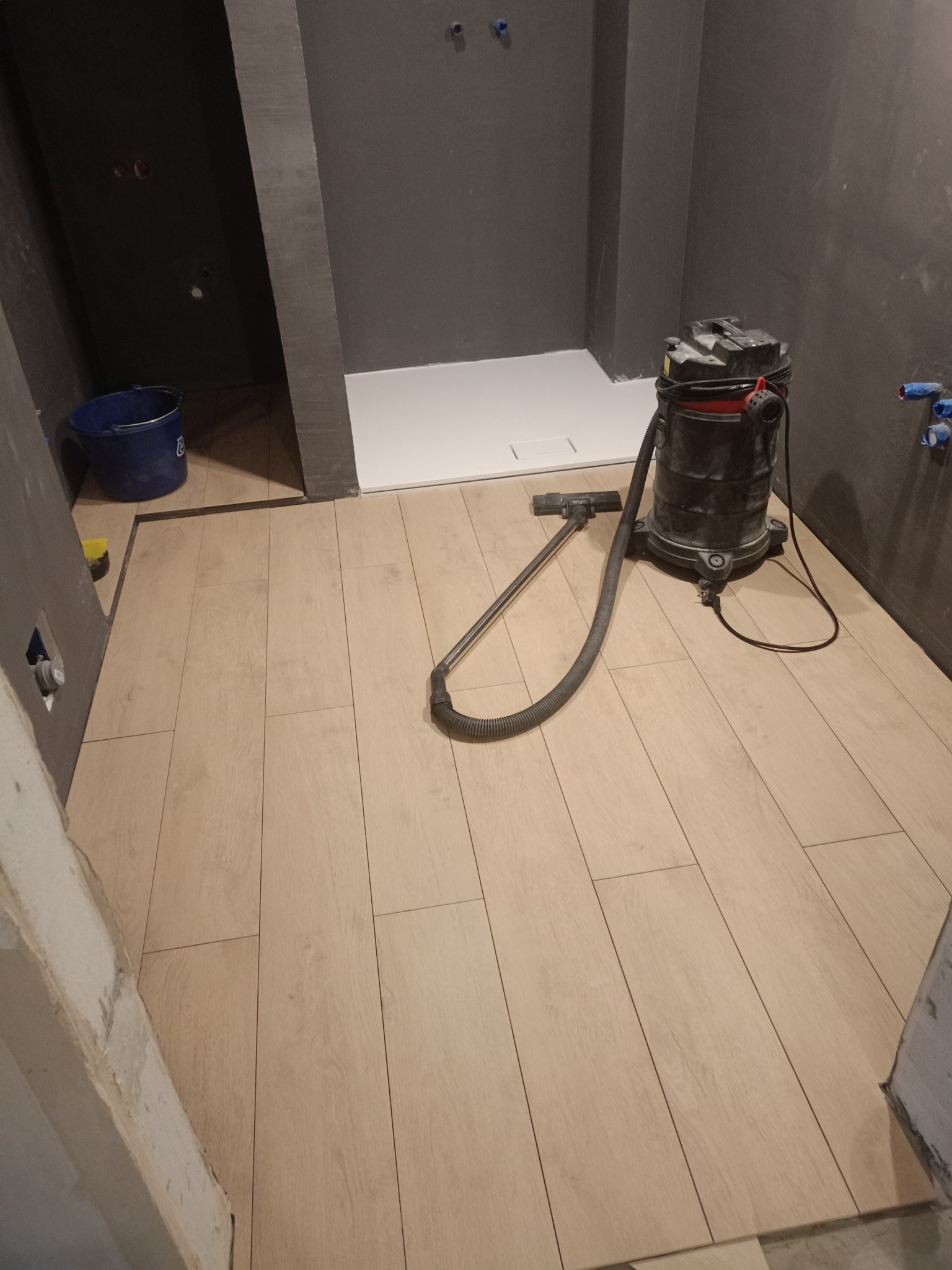Jasne panele podłogowe w łazience z szarą ścianą i brodzikiem. Odkurzacz przemysłowy stoi na świeżo ułożonej podłodze. Widoczne niedokończone wykończenia.