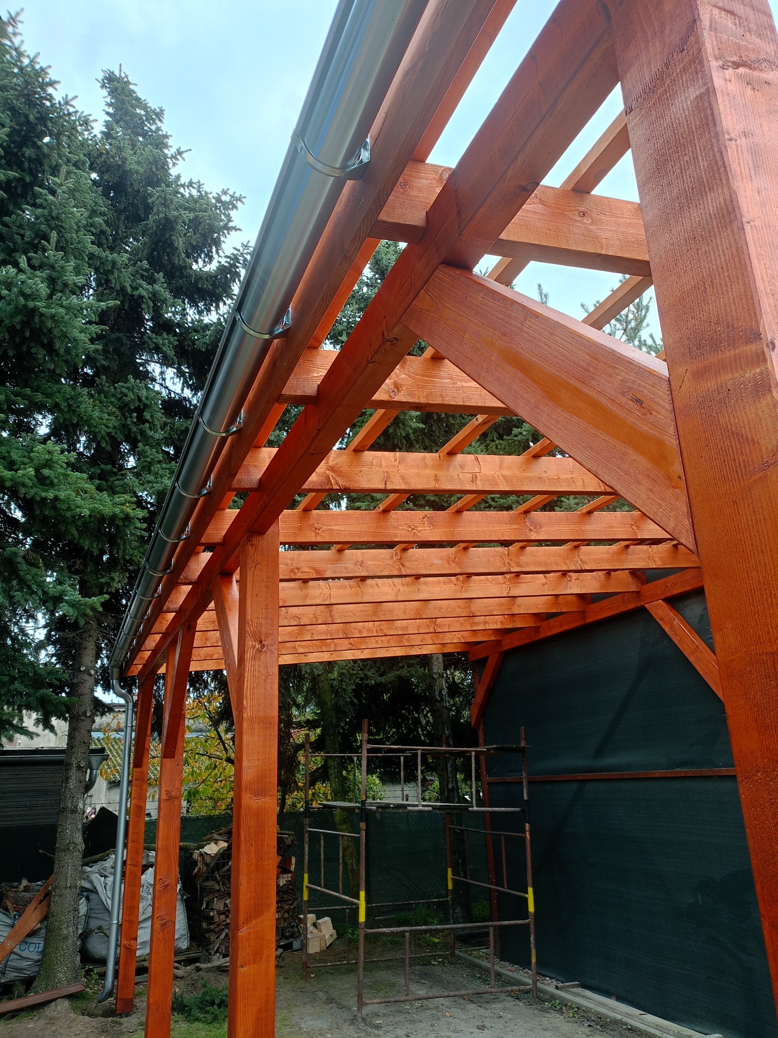 Drewniana konstrukcja z orynnowaniem, widok z dołu. Świeżo zaimpregnowane drewno w ciepłym odcieniu brązu. W tle drzewa i rusztowanie.