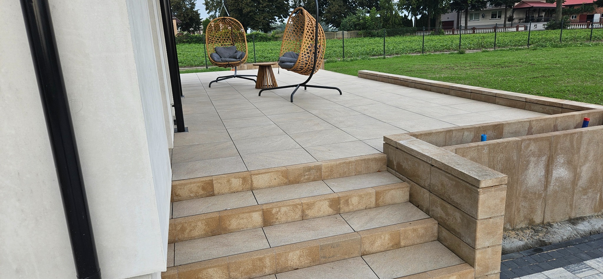 Jasne, kwadratowe płyty tarasowe tworzą schody i patio z dwoma wiszącymi fotelami. Widok na trawnik i domy w tle. Ujęcie z niskiego kąta.