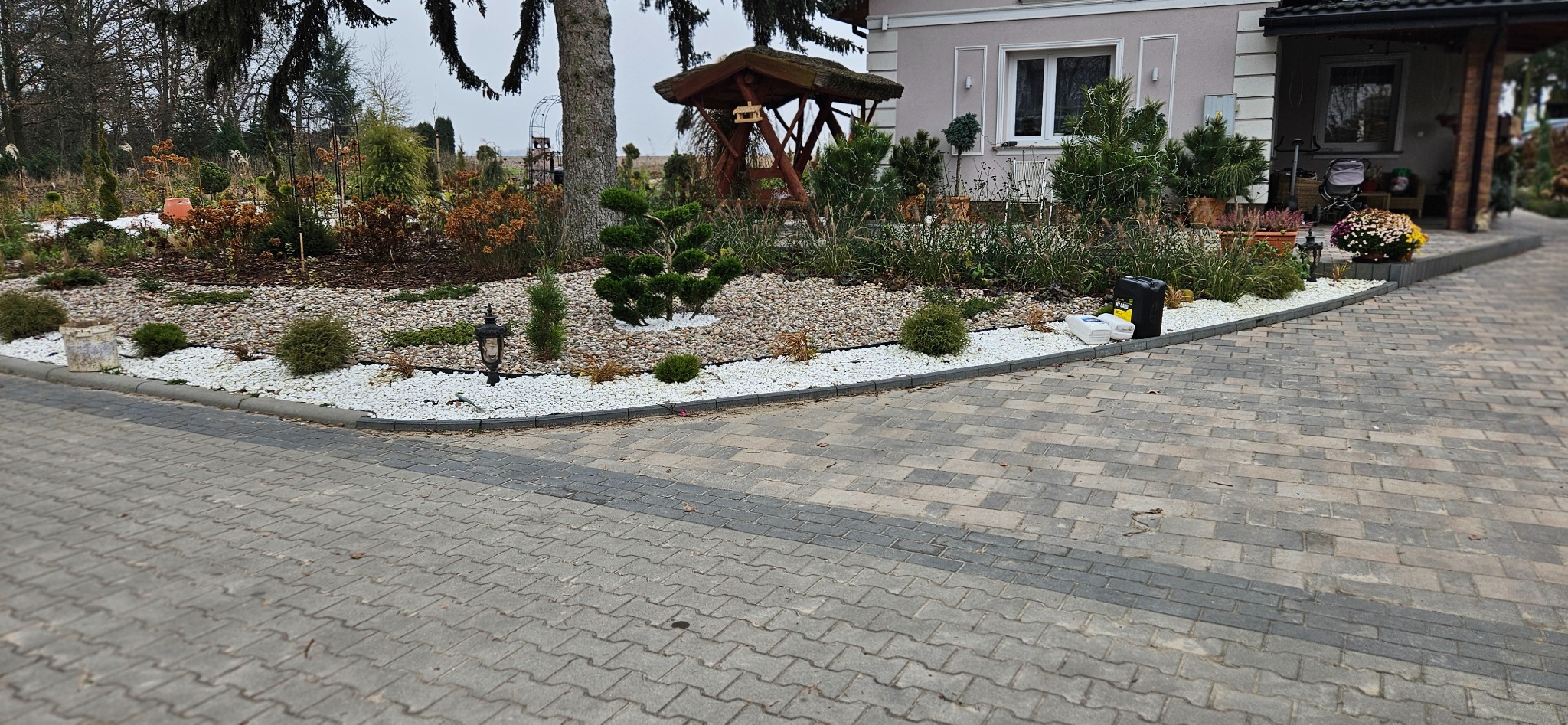 Ułożona kostka brukowa w szarych odcieniach, obrzeże z białych kamieni, rabata z roślinami i ozdobami, w tle dom jednorodzinny i altana.