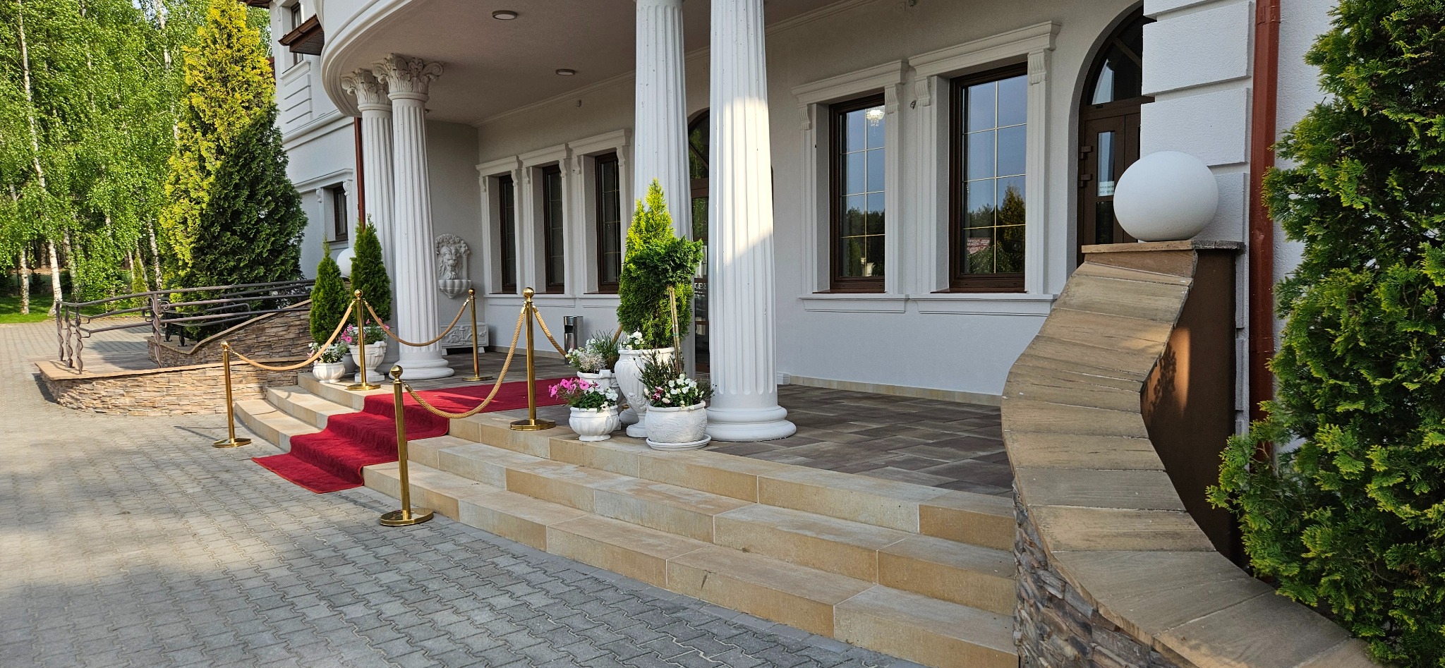 Eleganckie wejście do budynku z kolumnami, schodami z piaskowca, czerwonym dywanem i dekoracyjnymi roślinami w donicach. Kostka brukowa na podjeździe.