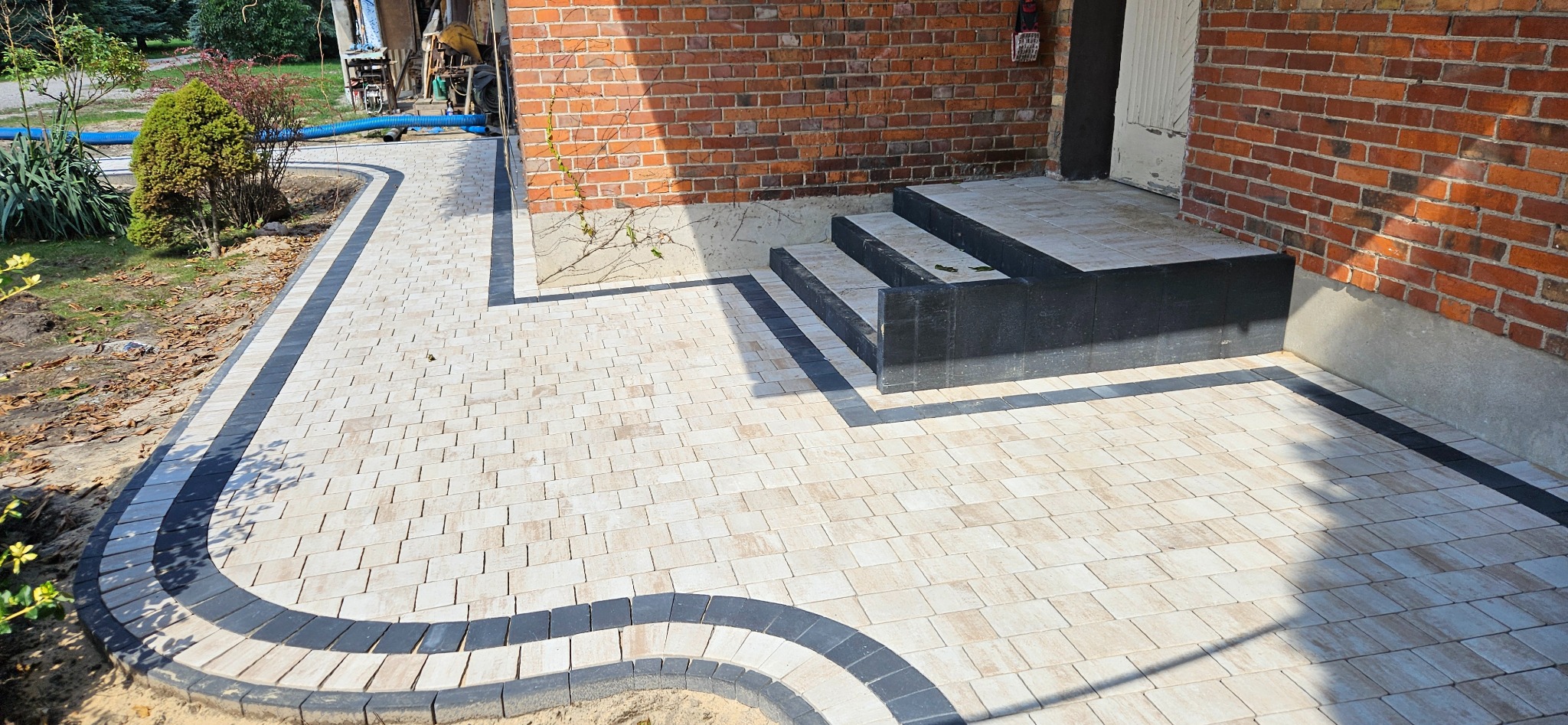 Jasna kostka brukowa z ciemnymi obrzeżami tworzy elegancki wzór ścieżki i schodów przy ceglanym budynku. Widoczny fragment ogrodu z roślinnością.