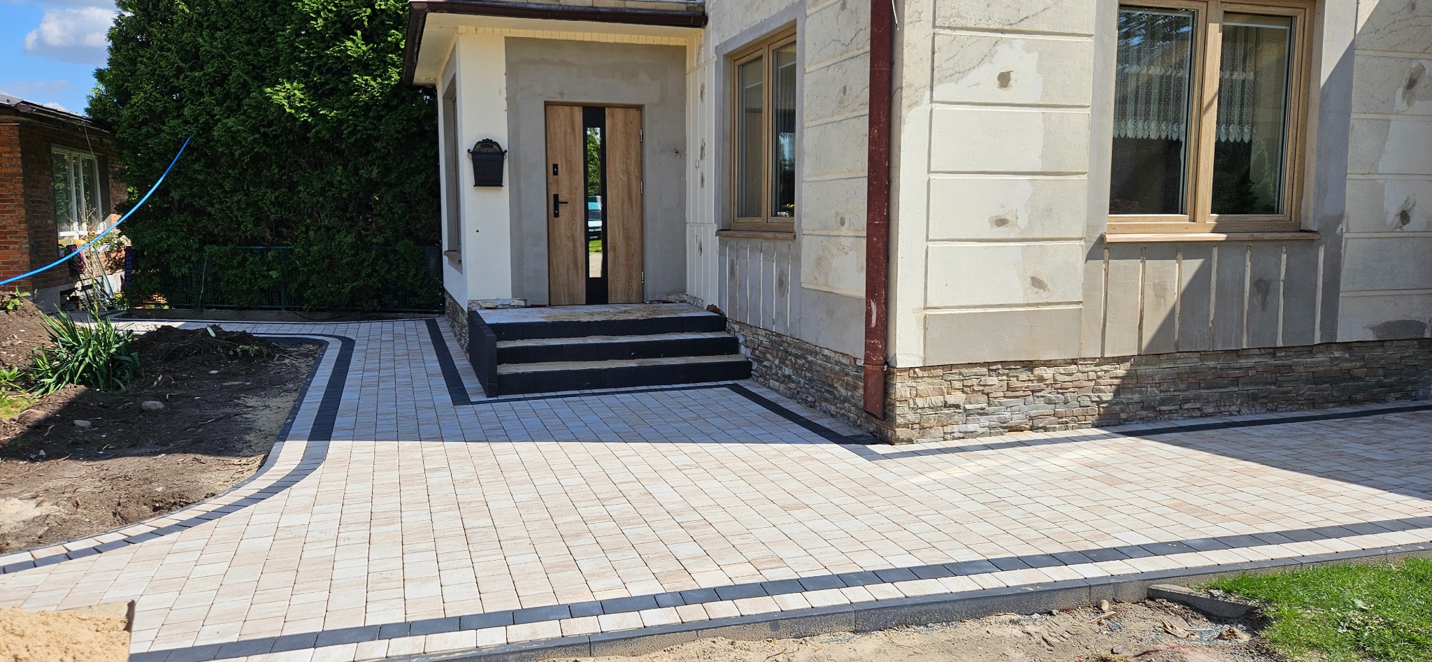 Jasna kostka brukowa z czarną obwódką prowadzi do wejścia domu z drewnianymi drzwiami. Widoczne okno i fragment elewacji z kamienną okładziną.