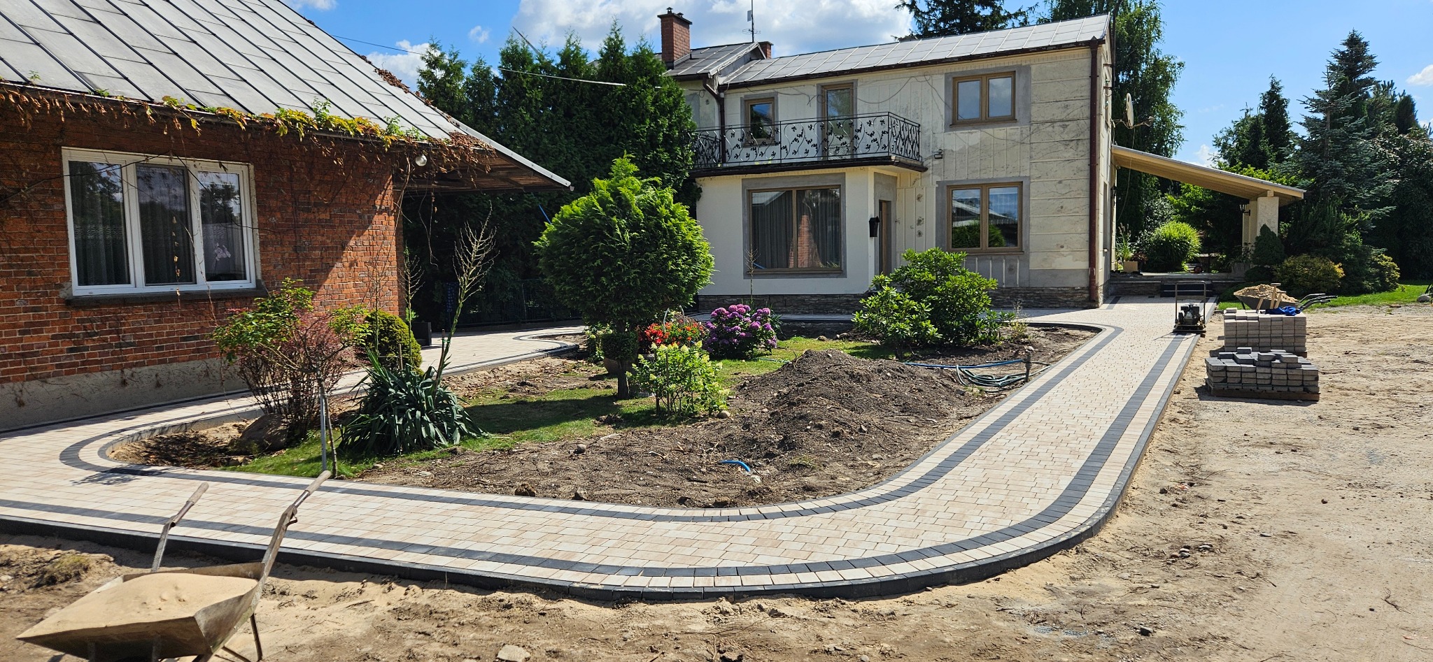 Ułożona ścieżka z kostki brukowej w ogrodzie, obrzeża z ciemnej kostki, w tle dom z balkonem i zieleń. Częściowo ułożona kostka, widoczna taczka i maszyna do ubijania.