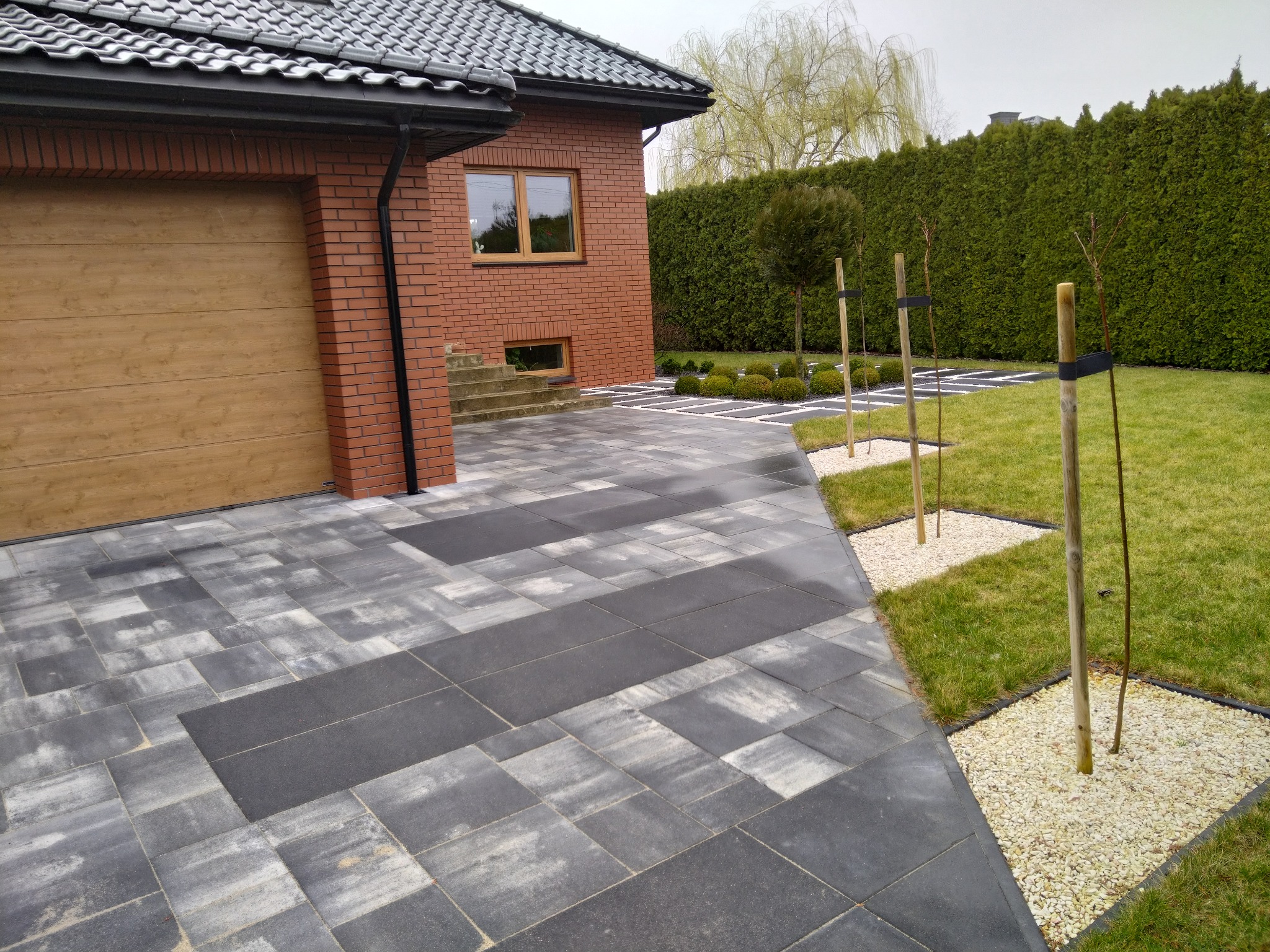 Nowoczesny podjazd z szarej kostki brukowej o geometrycznym wzorze, obok domu z cegły i zielonego trawnika z młodymi drzewkami. Widok na garaż i część ogrodu.