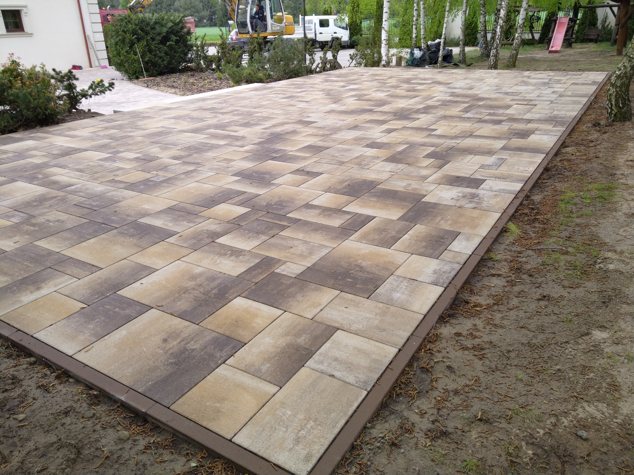 Ułożona kostka brukowa w odcieniach beżu i brązu tworzy dużą, prostokątną powierzchnię. W tle widoczny sprzęt budowlany i zieleń drzew.