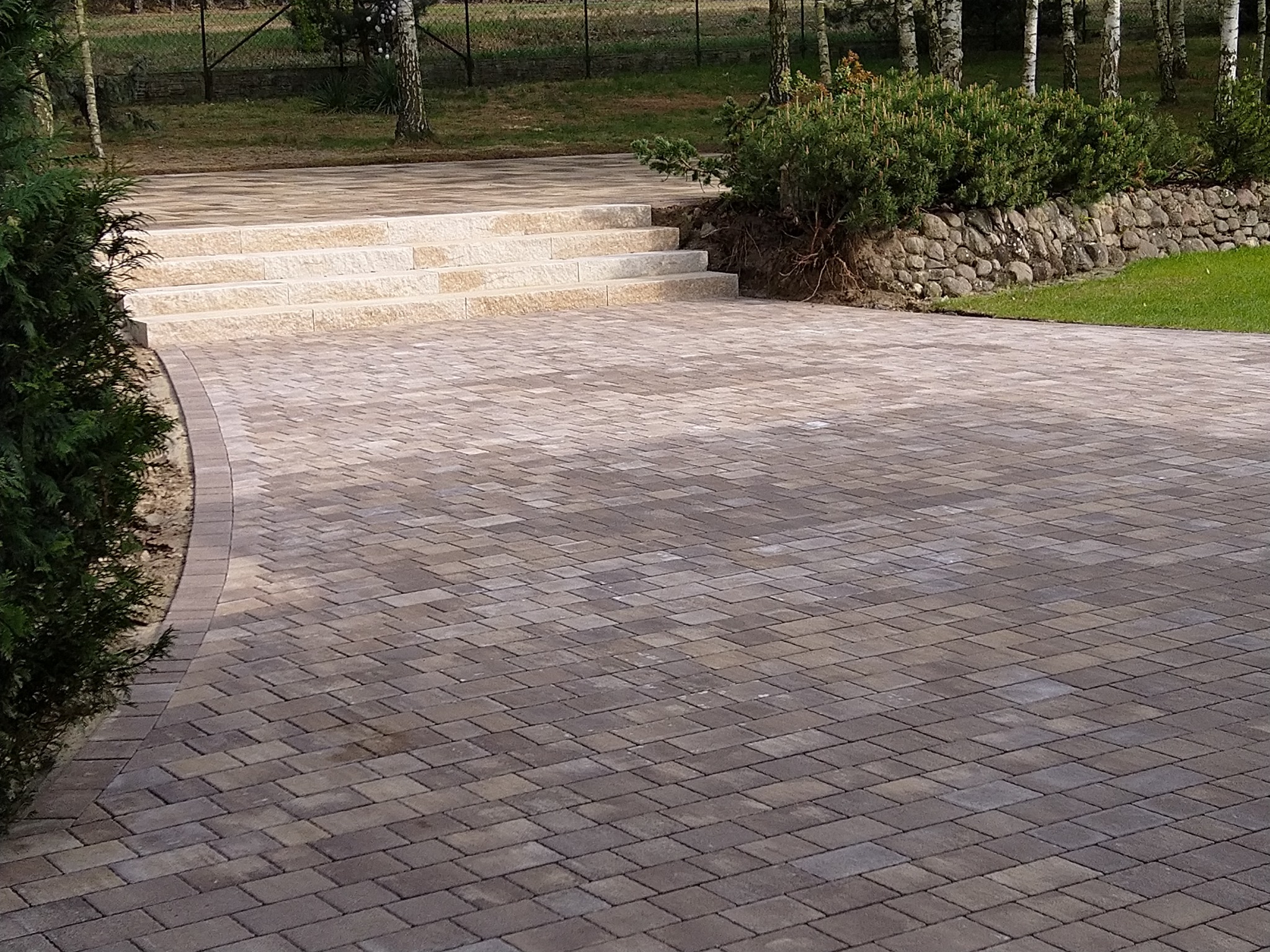 Ułożona kostka brukowa w odcieniach szarości i brązu tworzy ścieżkę z kamiennymi schodami i obrzeżem. Tło stanowią drzewa i roślinność.