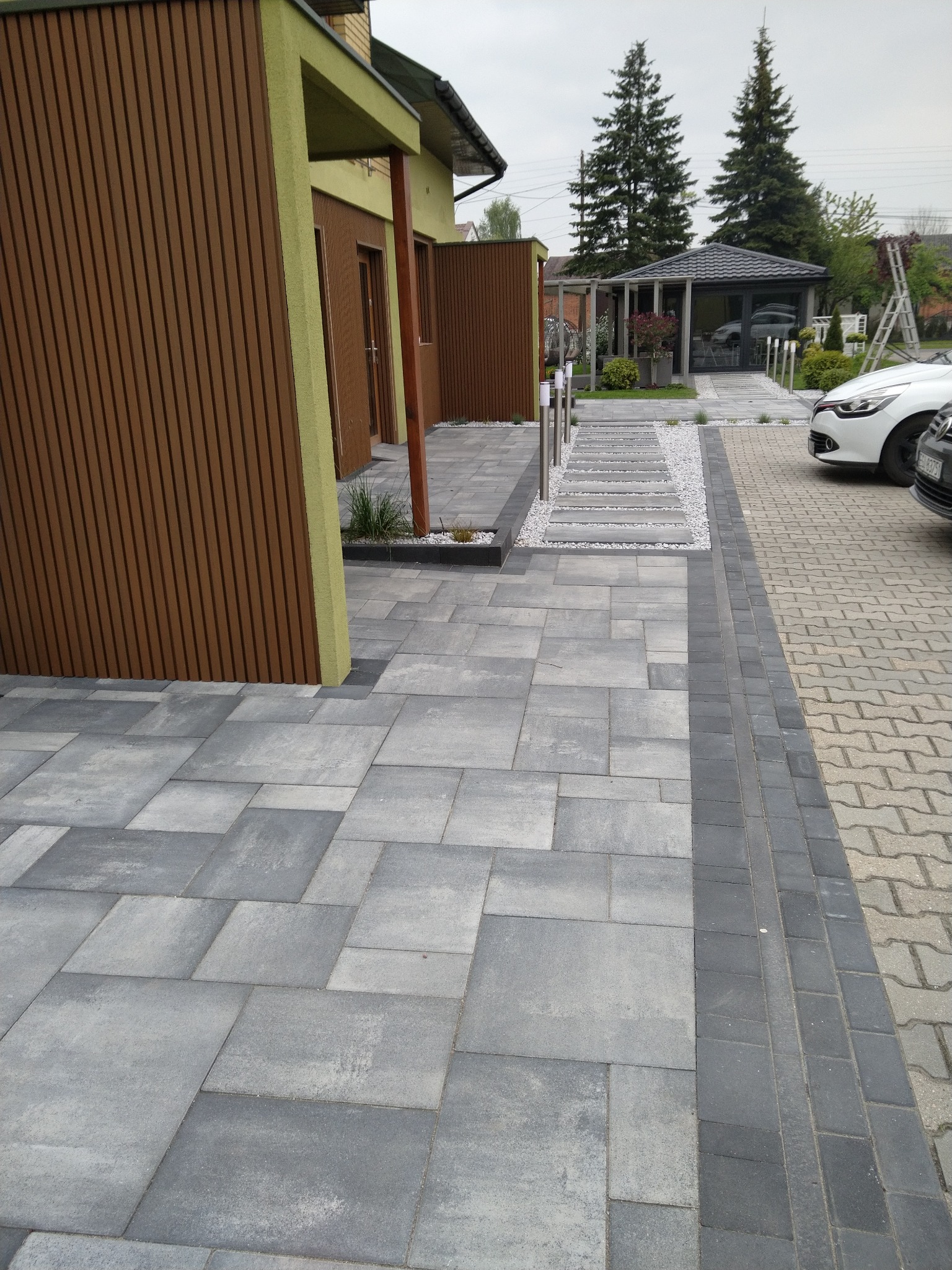 Ułożona szara kostka brukowa w różnych rozmiarach, obrzeża z ciemnej kostki. W tle budynek z drewnianą elewacją i ścieżka z płyt w otoczeniu białego żwiru.