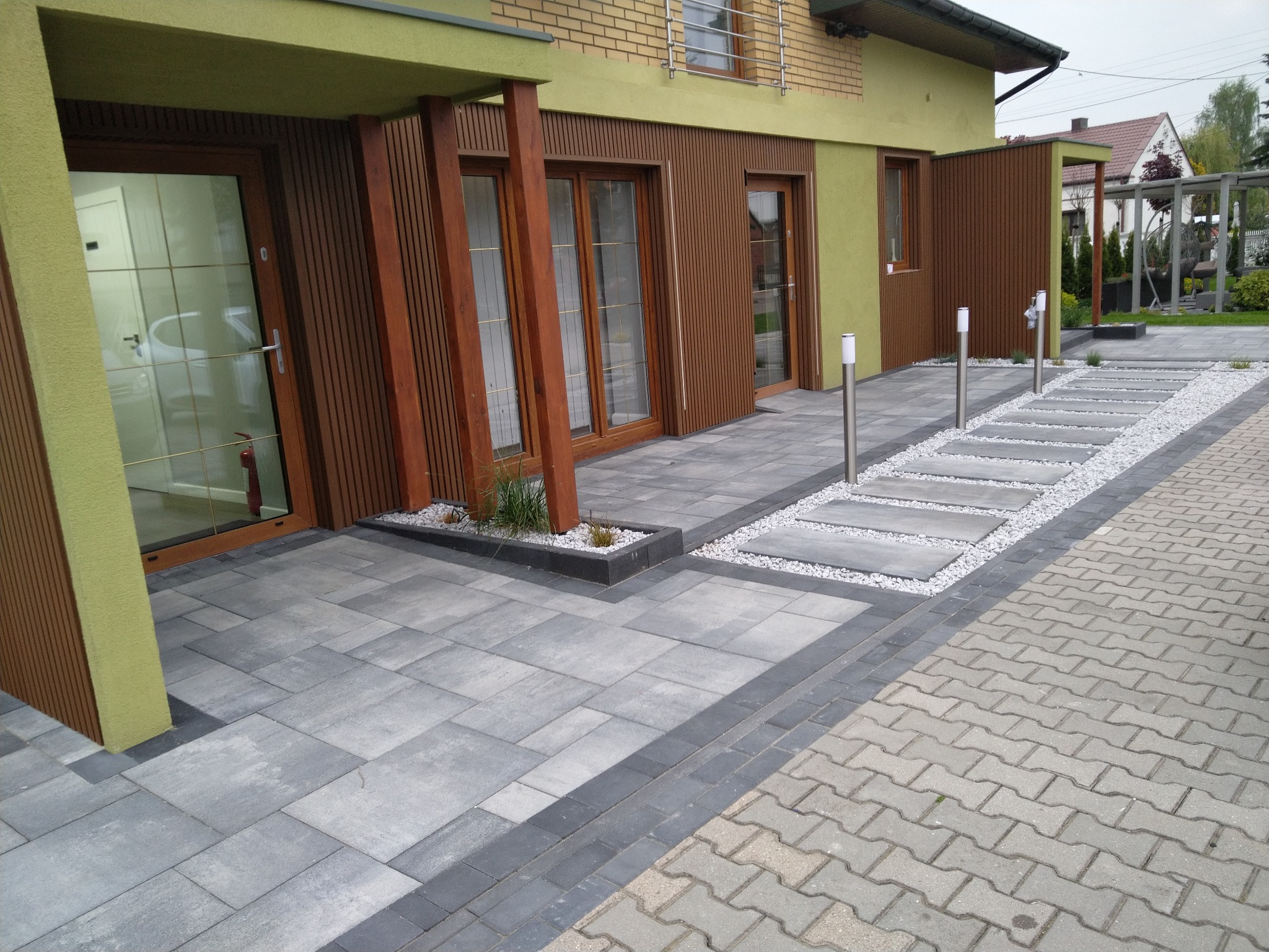 Nowoczesna aranżacja przestrzeni przed domem z szarej kostki brukowej, obrzeża z ciemnej kostki, ścieżka z płyt i białego grysu, oświetlenie punktowe.
