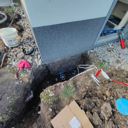 GeoPrecyzja - Wykop fundamentu budynku z widocznymi elementami instalacji podziemnej, narzędziami i materiałami budowlanymi na ziemi. Ujęcie z góry, w trakcie prac ziemnych.