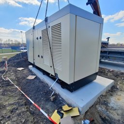 GeoPrecyzja - Montaż agregatu prądotwórczego na betonowym fundamencie za pomocą dźwigu. Widoczne łańcuchy, teren budowy zabezpieczony taśmą.