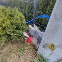 GeoPrecyzja - Wytyczenie trasy kabla w ziemi, oznaczone różowym palikiem i białą taśmą, przy słupku ogrodzeniowym z tabliczką ostrzegawczą. Widoczny niebieski kabel w peszlu.