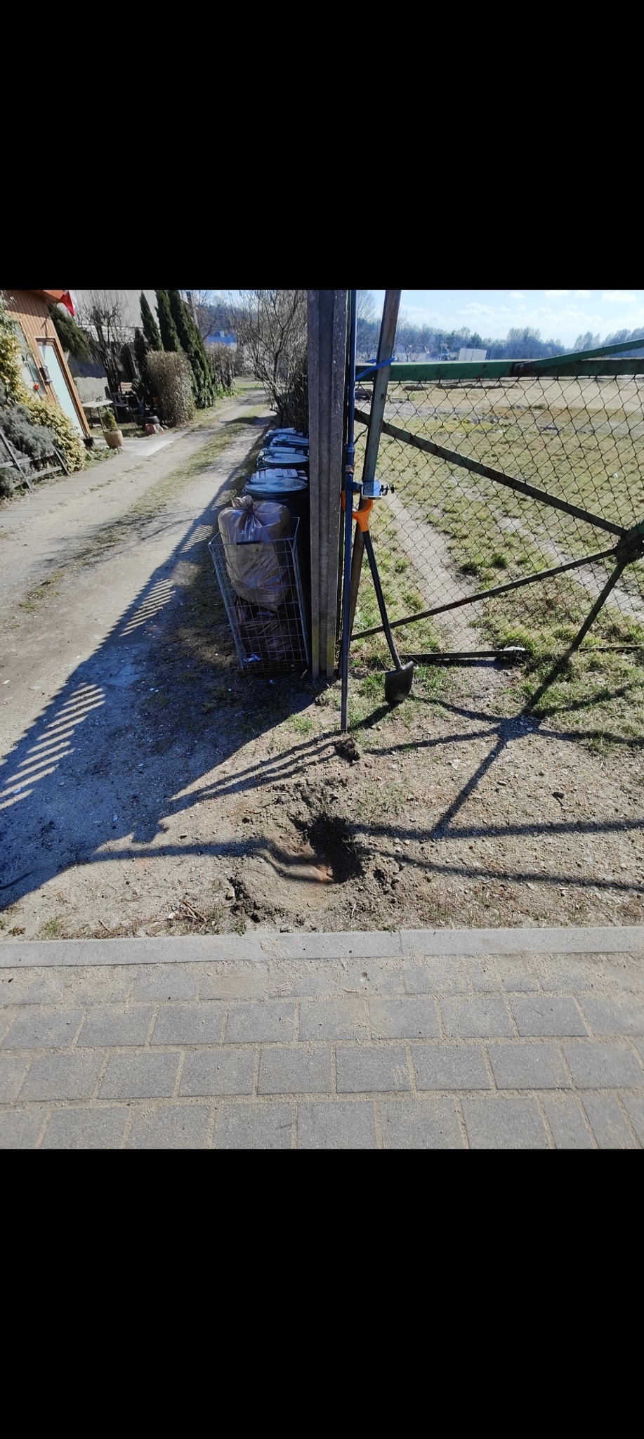 Wykonane pomiary gruntu przy ogrodzeniu, widoczny wykop i narzędzia pomiarowe. W tle worki na śmieci i zabudowa jednorodzinna.