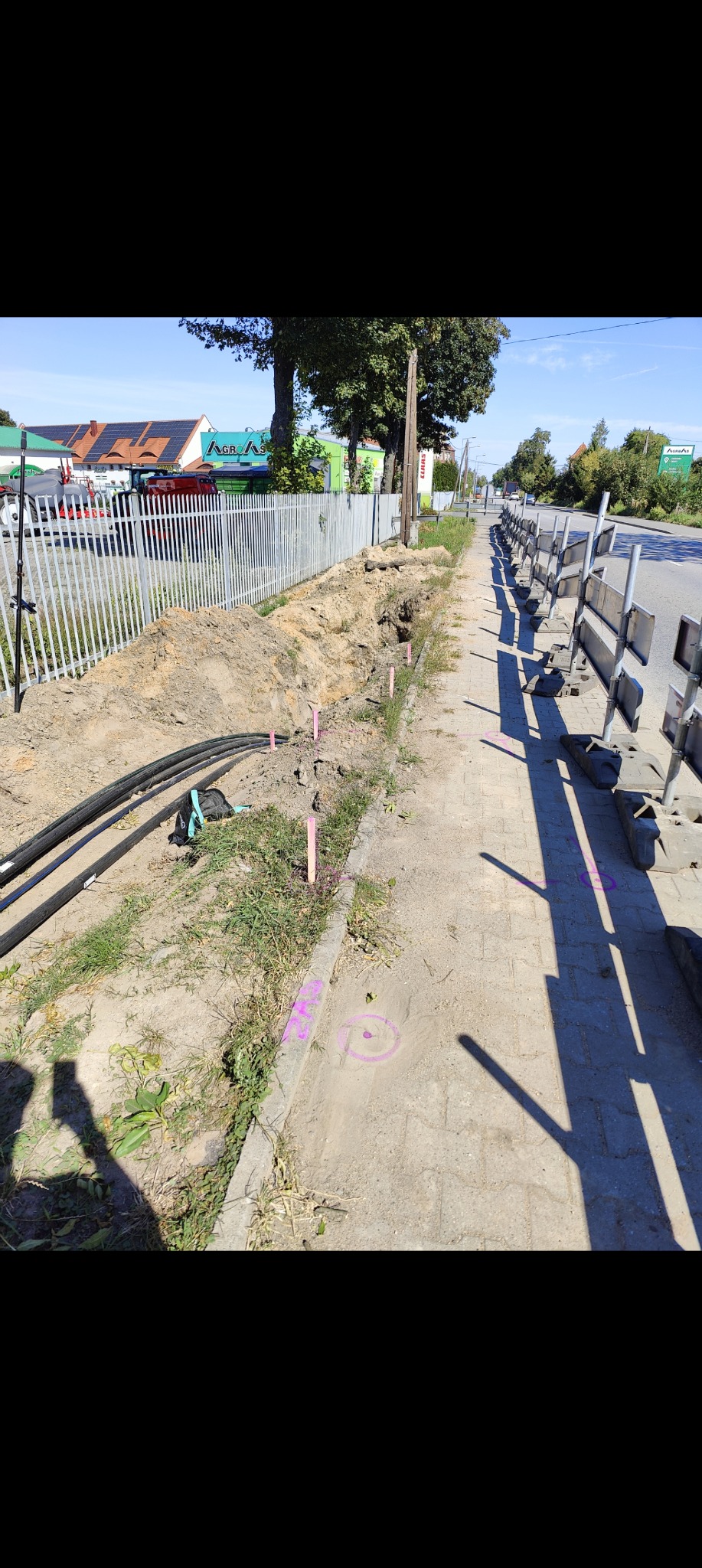 Prace ziemne przy chodniku z widocznymi rurami i oznaczeniami geodezyjnymi w postaci różowych palików i okręgów. W tle budynki i zieleń.