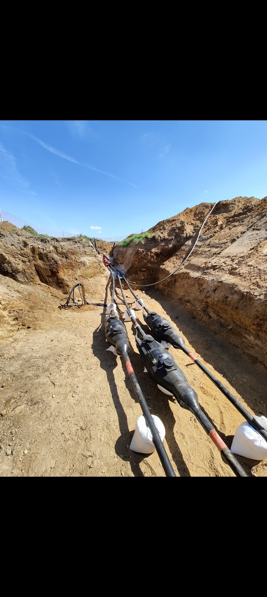 Kable energetyczne ułożone w wykopie, zabezpieczone workami. Widok z góry na instalację kablową w ziemi, błękitne niebo w tle. Prace ziemne.