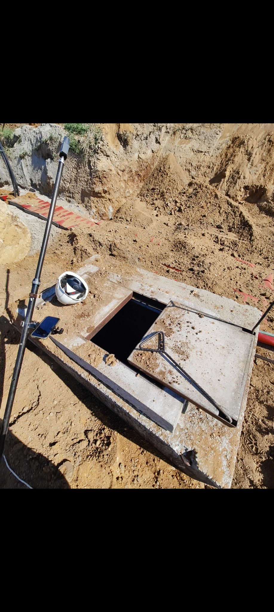 Otwarty właz betonowy w wykopie, obok biały kask i sprzęt pomiarowy. Widoczne kable i ziemia. Prace ziemne w toku, teren budowy.