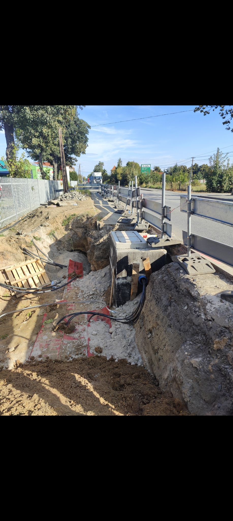 Wykop z zabezpieczeniami przy drodze, widoczne kable i skrzynka betonowa. Paleta drewniana i oznaczenia geodezyjne na dnie wykopu. Prace ziemne w toku.