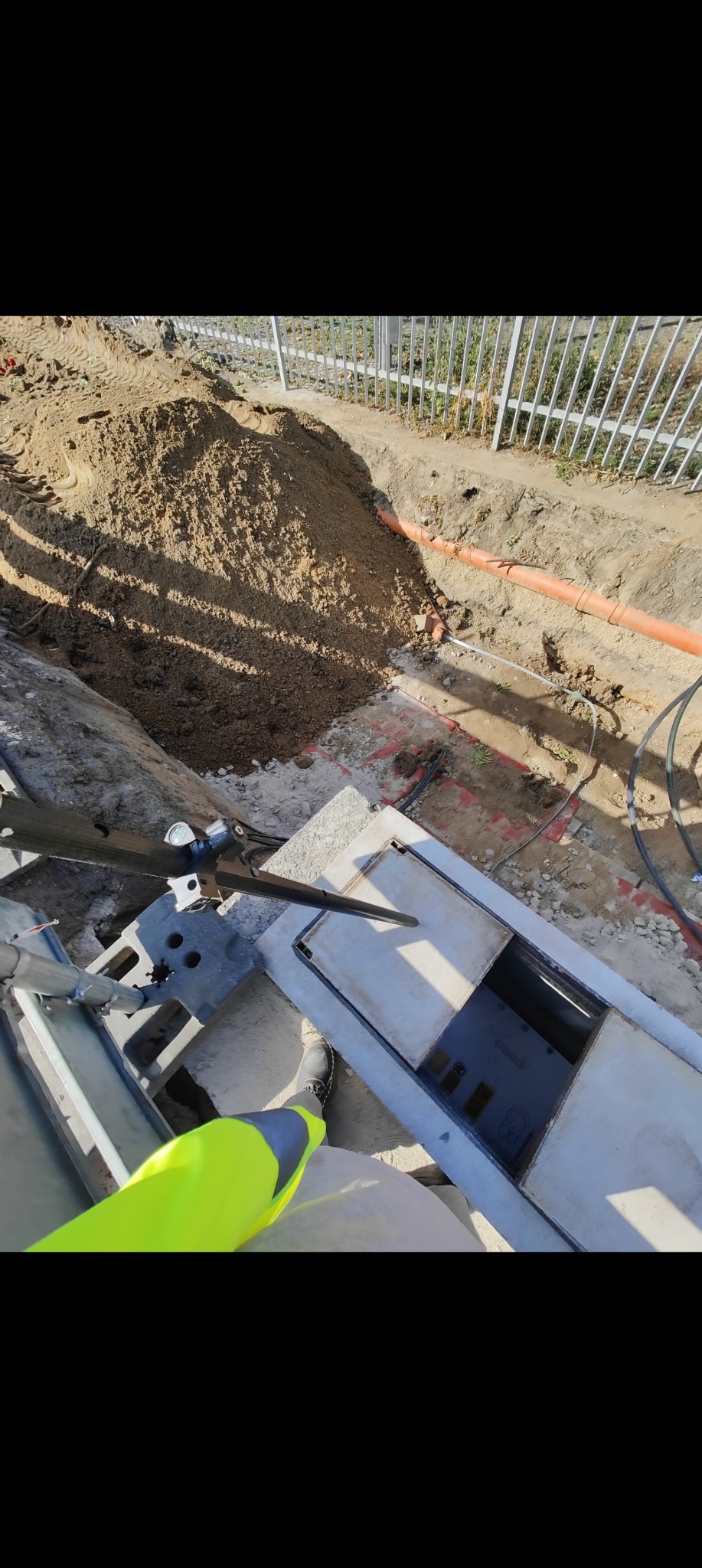 Prace geodezyjne w terenie: wykop z pomarańczowymi rurami, studzienka betonowa z otwartą pokrywą, sprzęt pomiarowy i osoba w odblaskowej kamizelce.