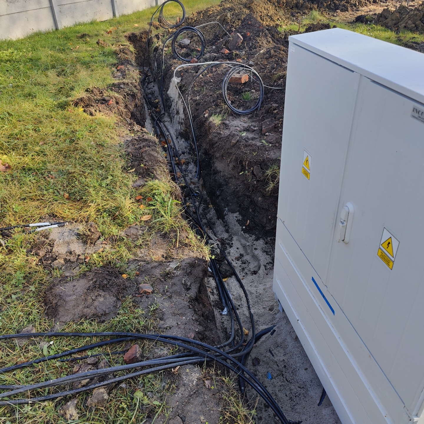 Wykop z ułożonymi kablami telekomunikacyjnymi, obok szafa sterownicza. Widoczne elementy instalacji podziemnej na trawiastym terenie.